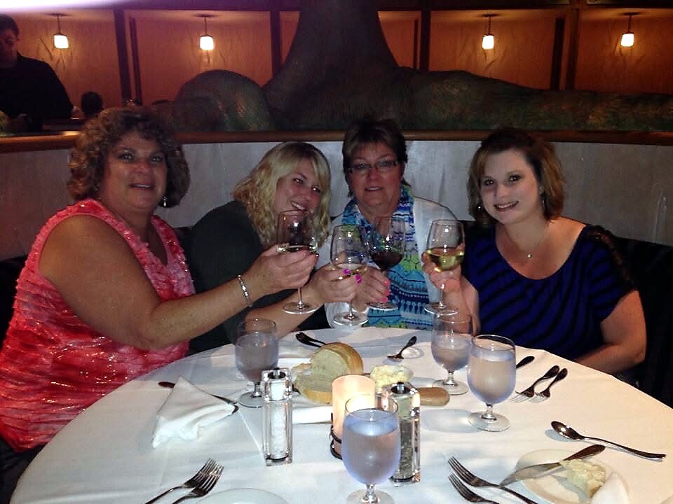Group of four women toasting with wine glasses at a restaurant table, smiling and dressed up for dinner.