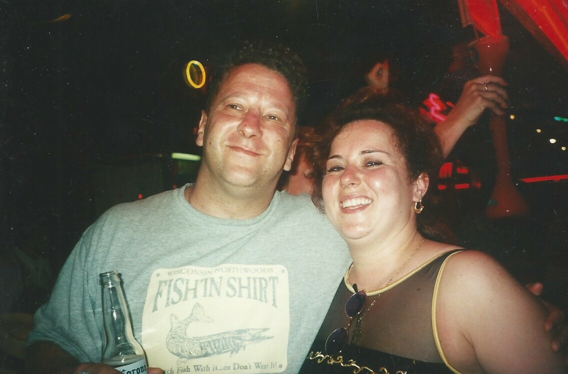 Two friends at a party pose for a photo, man holding a beer bottle and woman smiling beside him in a club setting.