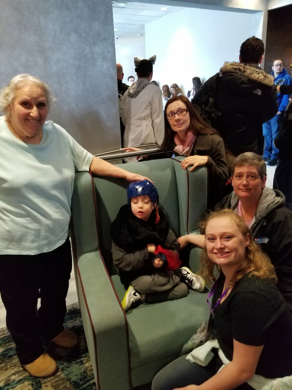 Child in a blue helmet sits on a green sofa, surrounded by smiling adults in a busy indoor lobby.