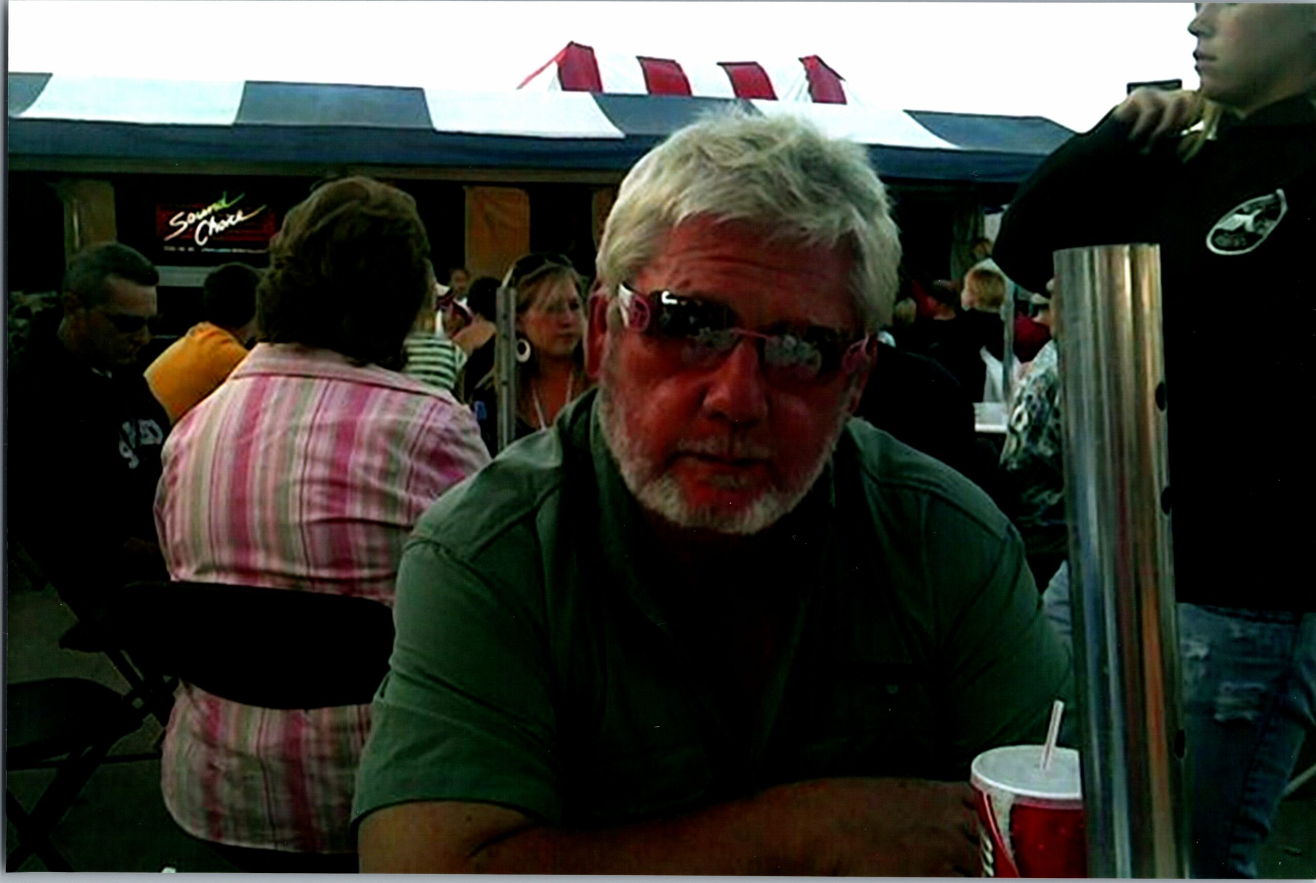 Man with gray hair and beard wearing sunglasses sits at an outdoor cafe table; a red cup with straw is on the table in front of him, with patrons in the background