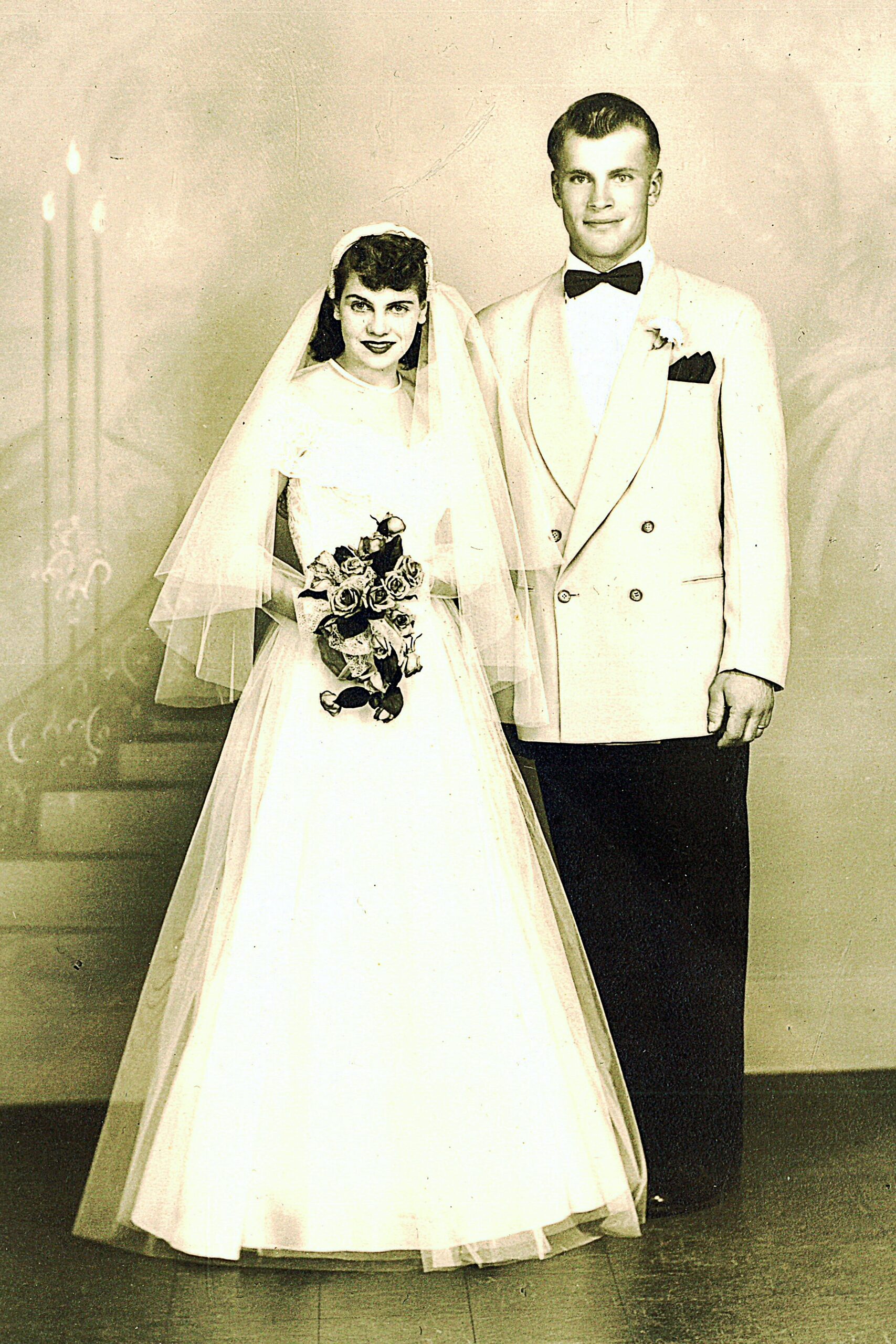 Vintage wedding photo: bride in white gown with veil holding a bouquet, standing next to a groom in a light suit with a black bow tie.