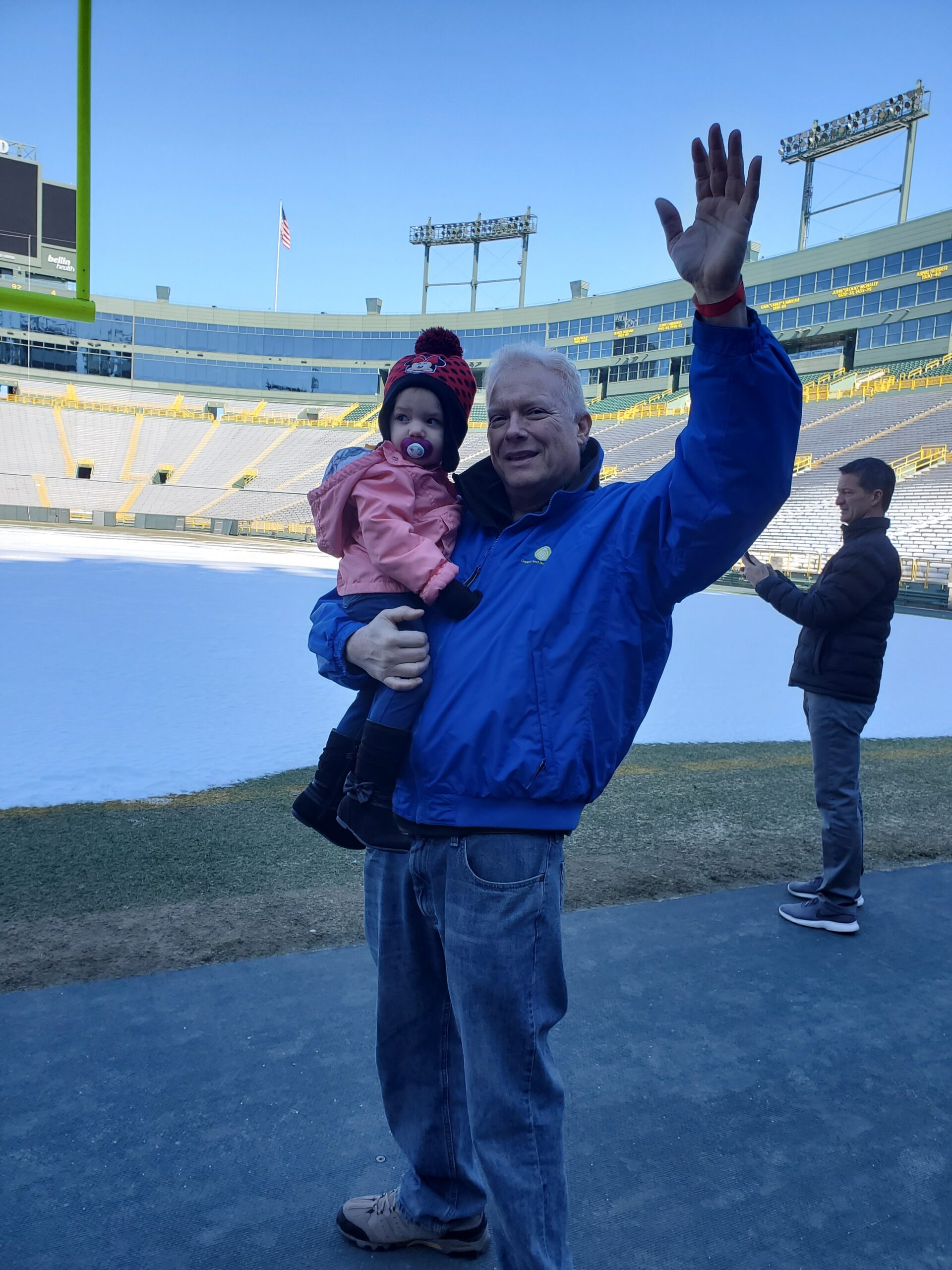 Man in blue jacket waving while holding a child at an empty football stadium rink backdrop behind them