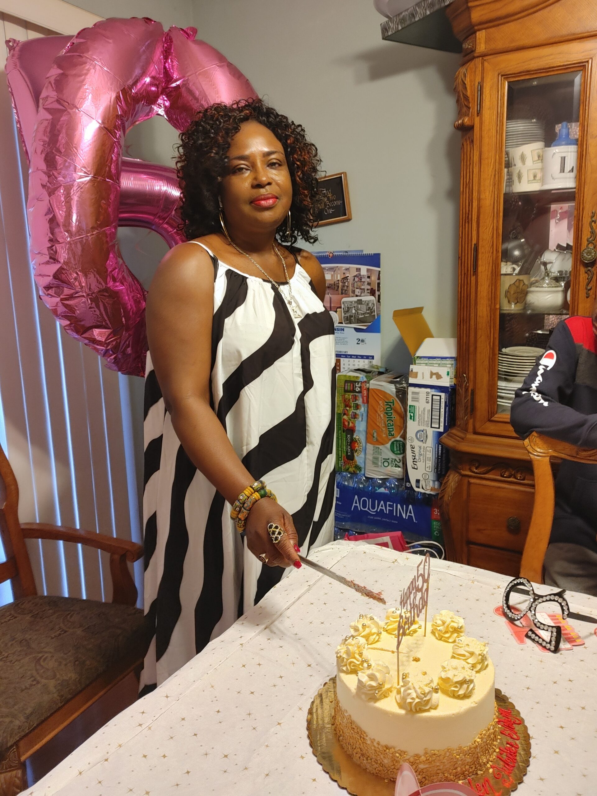 Woman in a black-and-white striped dress stands by a frosted cake with rosettes, pink foil balloon behind her.
