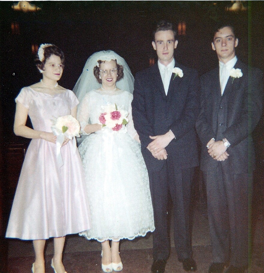 Bride in a white wedding gown with veil and bouquet, flanked by a bridesmaid in pink and two groomsmen in dark suits with boutonnieres.