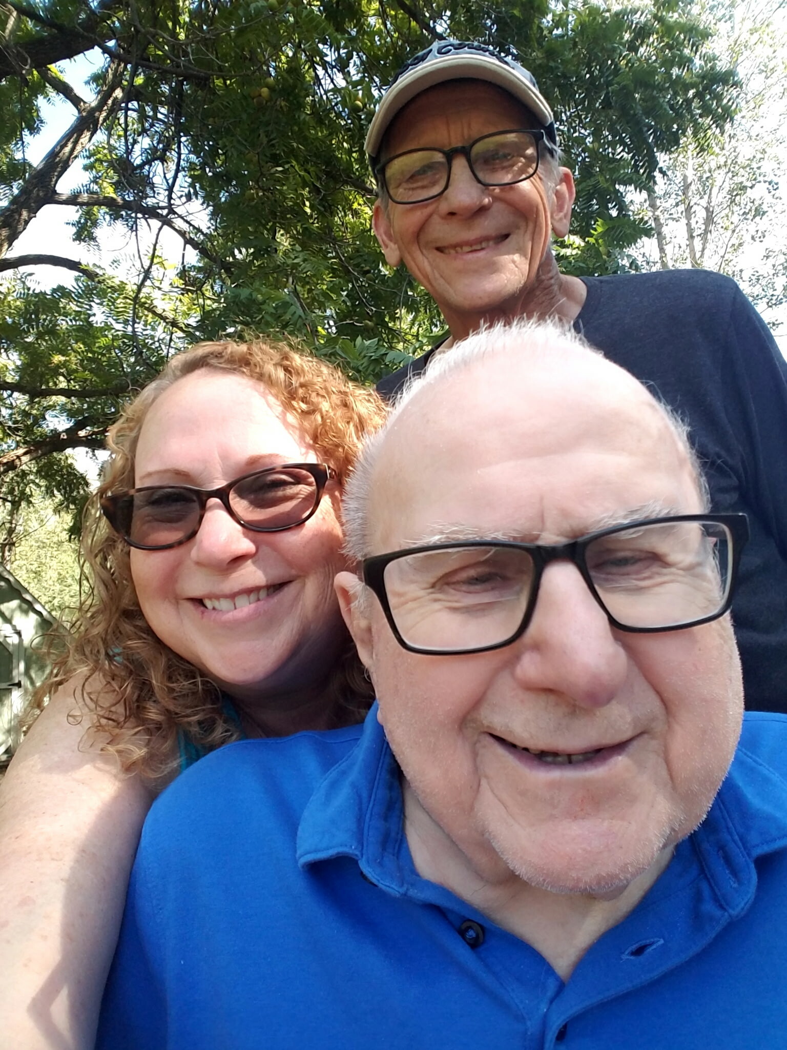 Three friends take a sunny outdoor selfie among green trees, smiling at the camera.