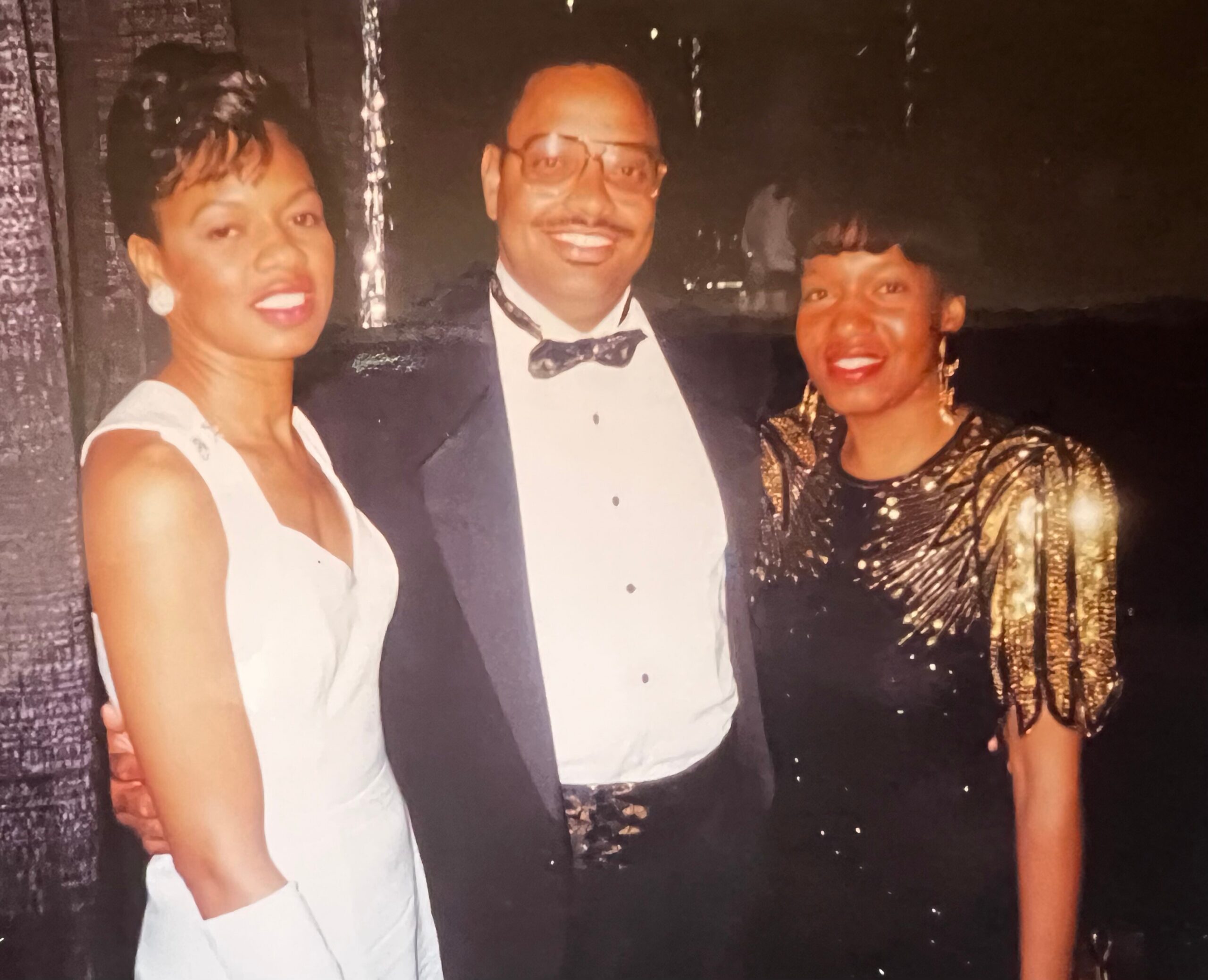 Three adults in formal wear posing for a group photo: woman in white on the left, man in a tuxedo center, woman in a gold dress on the right.