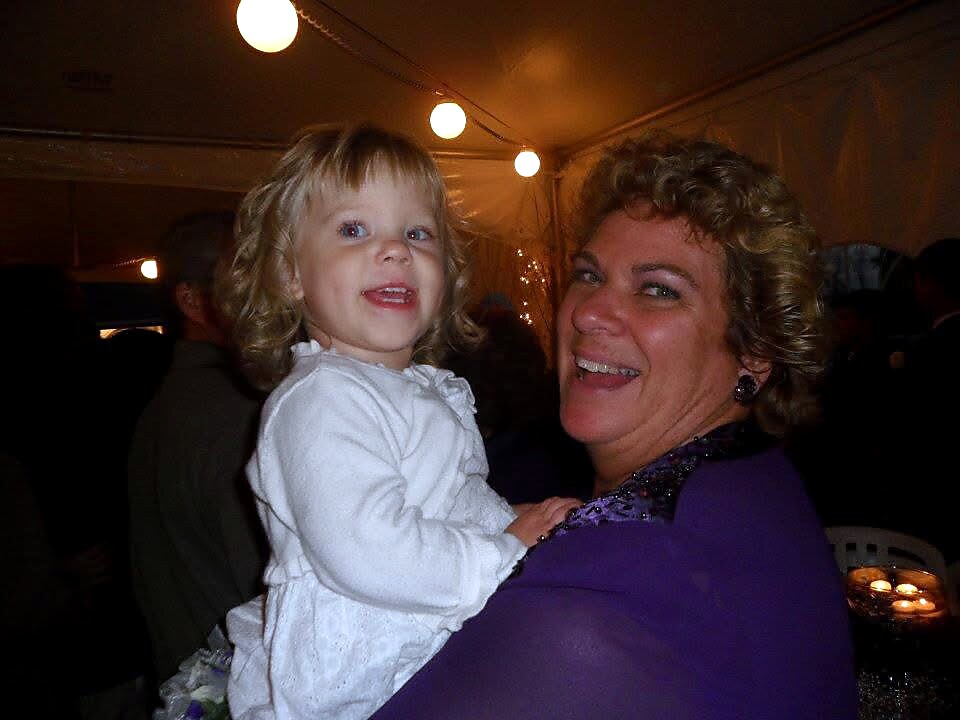 Woman in a purple outfit holds a smiling young girl in a white dress at a festive event under string lights in a tented venue.