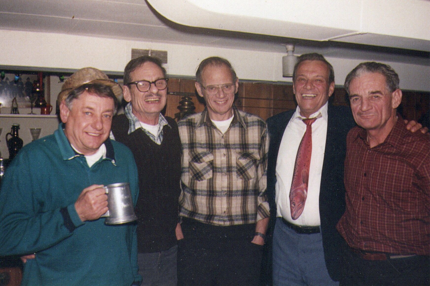 Five men posing together and smiling in a casual indoor setting; one holds a metal mug in the foreground.