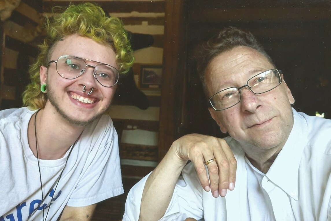 Two men pose for a selfie: a younger man with curly blond hair, glasses, a nose ring, and green ear gauge smiling next to an older man with glasses and a wedding ring, resting his chin on his hand.