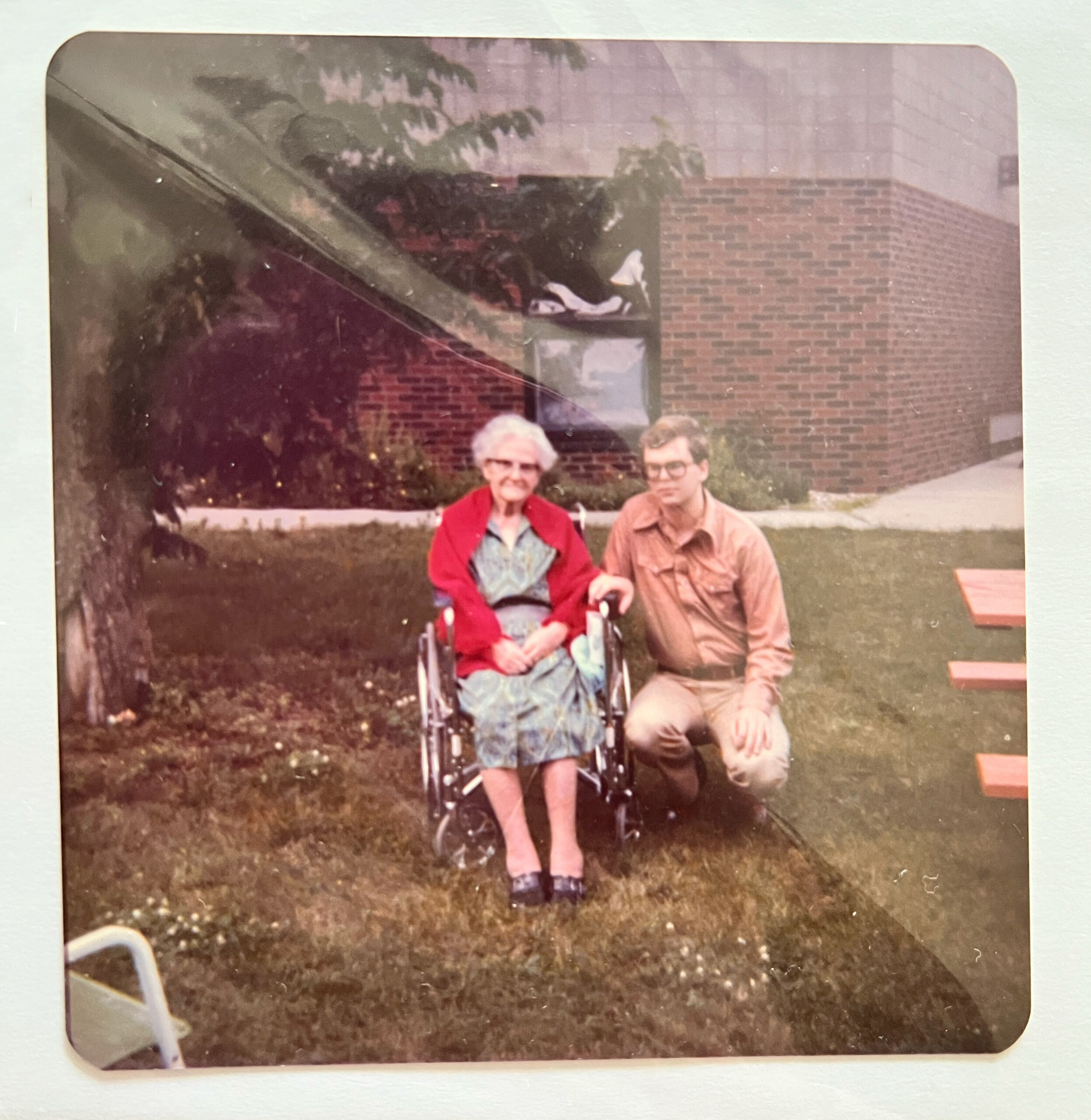 Two elderly people posing outdoors on a grassy area, one in a red jacket in a wheelchair beside a man in a tan shirt, with a brick building in the background.
