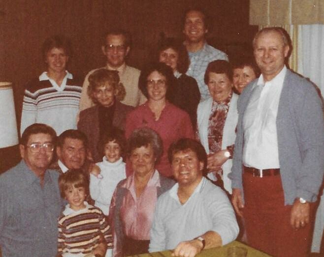 Large multi-generational family group poses indoors at a gathering; adults and children in casual outfits smiling for the camera, front row seated.