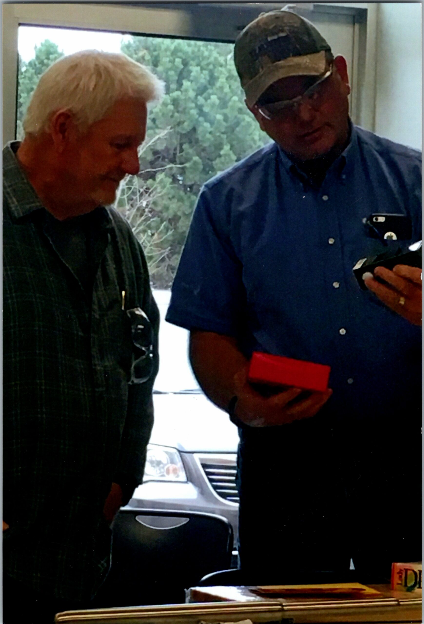 Two men indoors by a window; one with white hair in a plaid shirt, the other in a blue shirt and cap holding a red box.