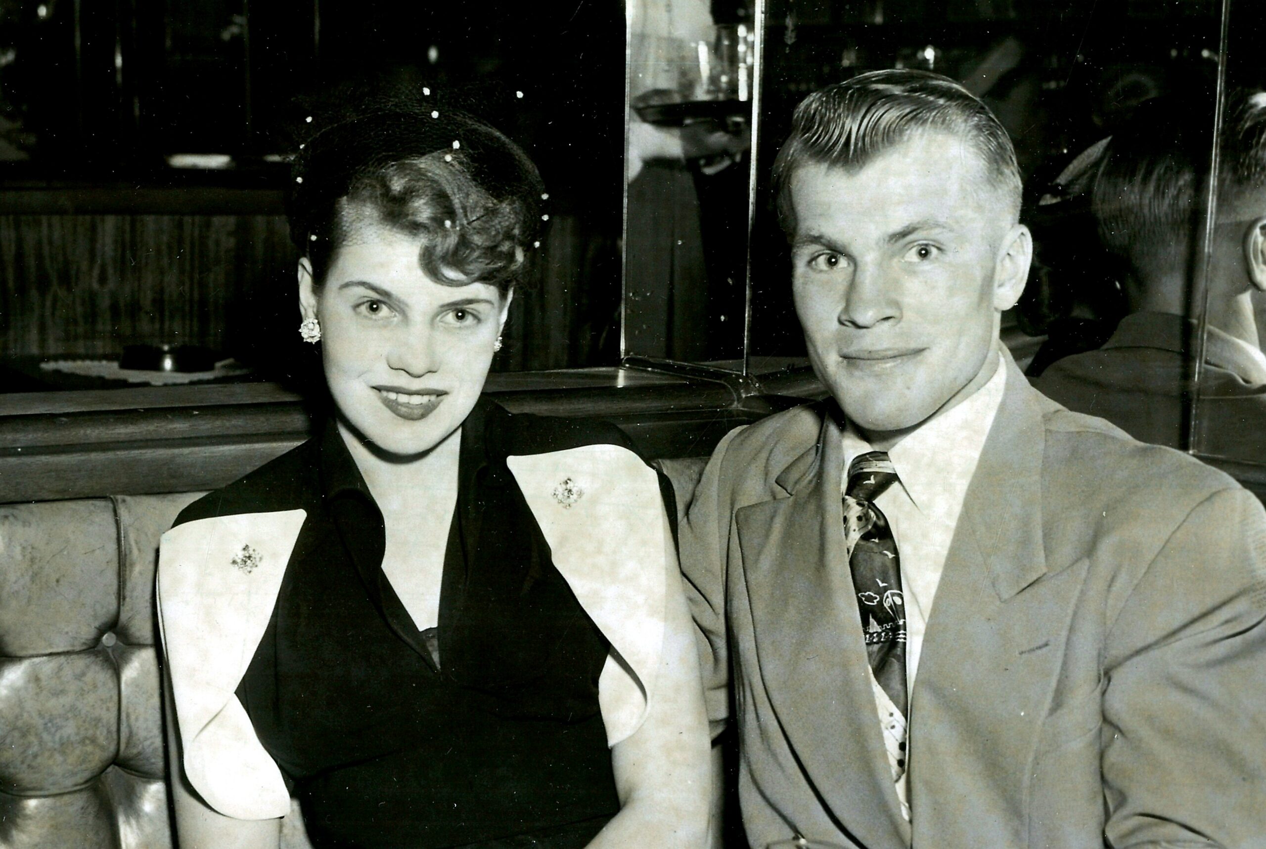 Black-and-white photo of a smiling couple in the backseat of a vintage car, dressed in 1950s attire; woman on left, man on right.