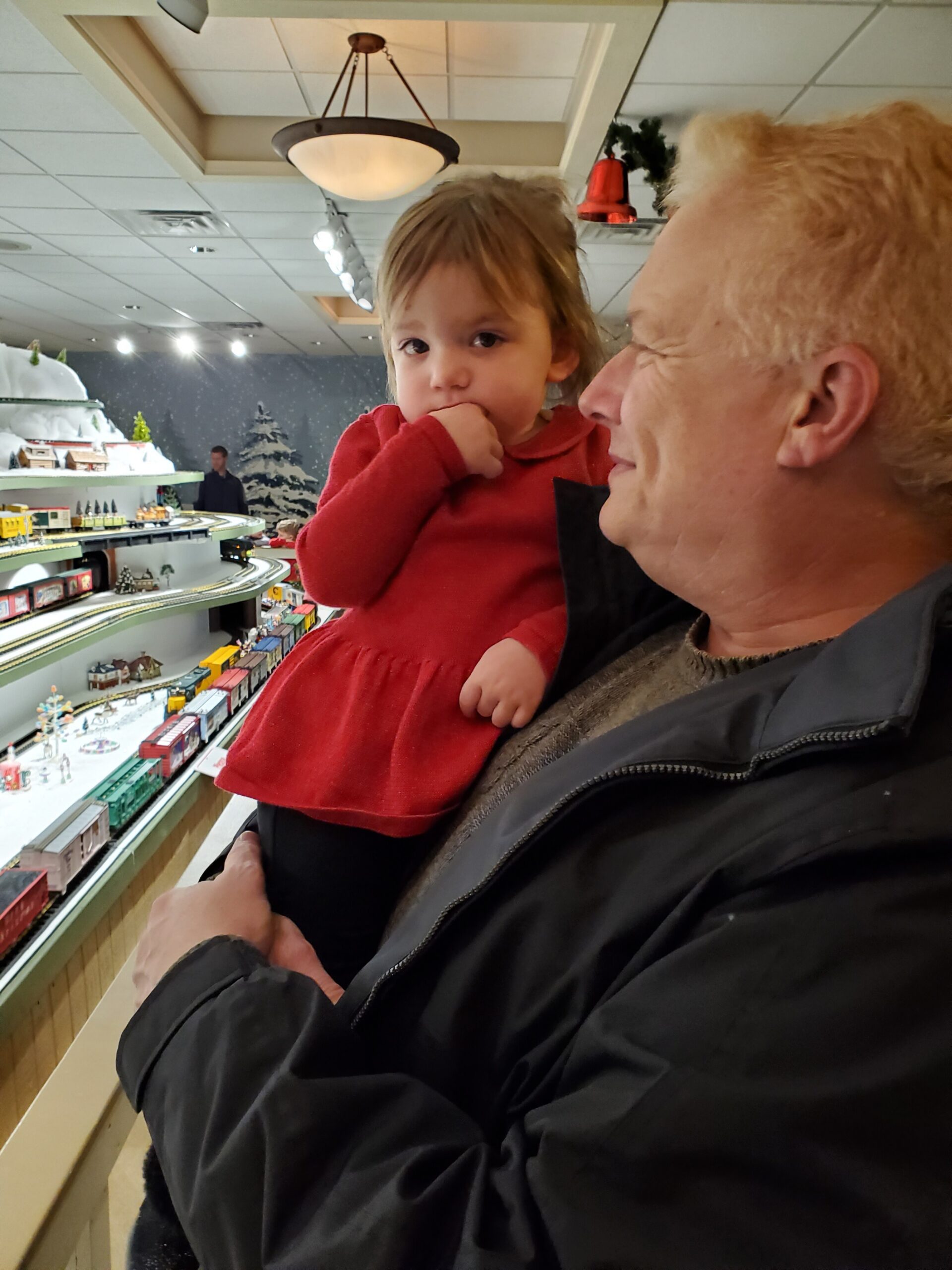 A man holds a young child in a red dress while looking at a large model train display in a store or exhibition hall.