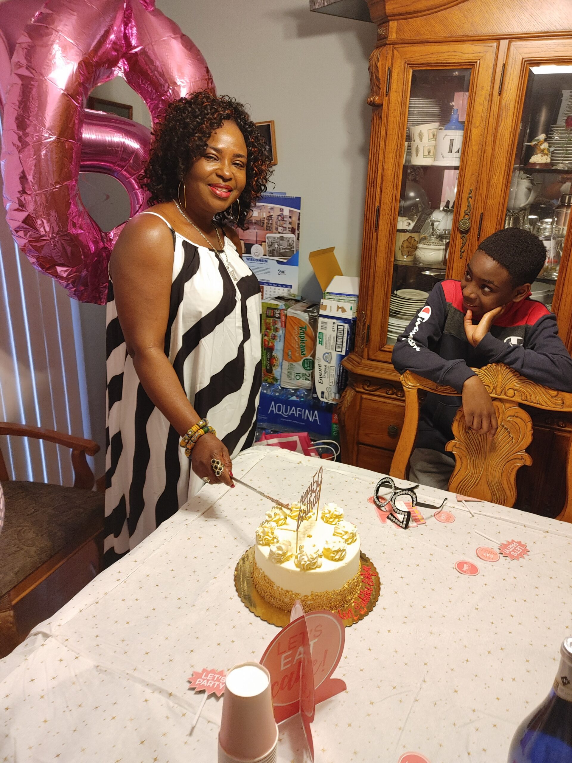 Woman in a black-and-white dress cuts a birthday cake at a table while a boy watches, with a pink number balloon in the background.