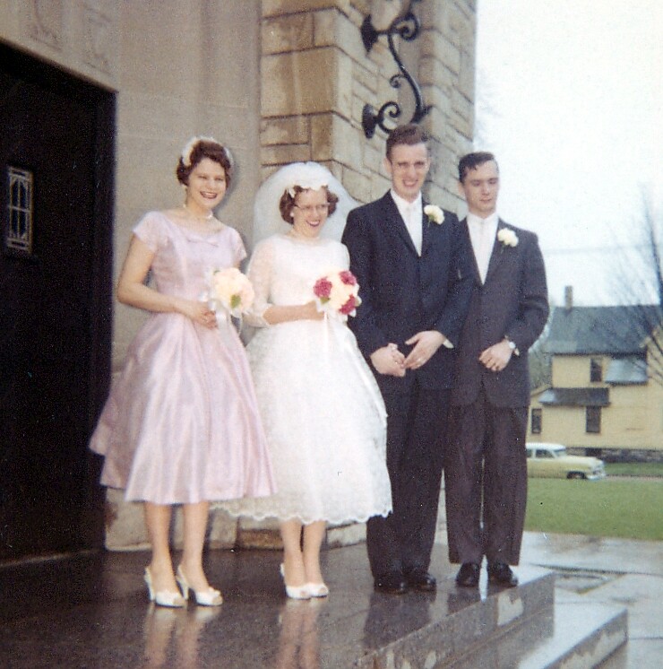 Bride in a white gown with a bouquet stands beside a bridesmaid in a pink dress and two men in dark suits outside a stone building, posing on steps.