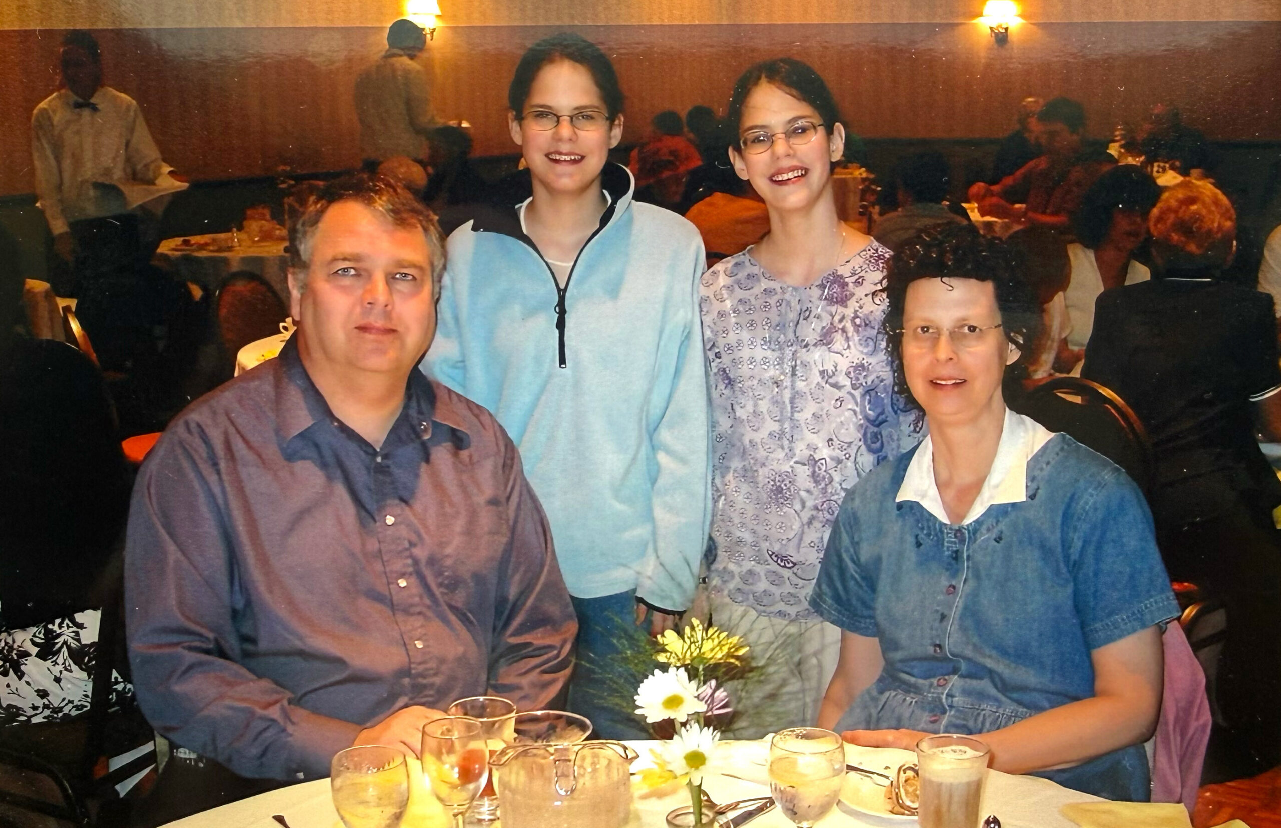Family of four posing at a restaurant table with drinks and a small flower centerpiece.