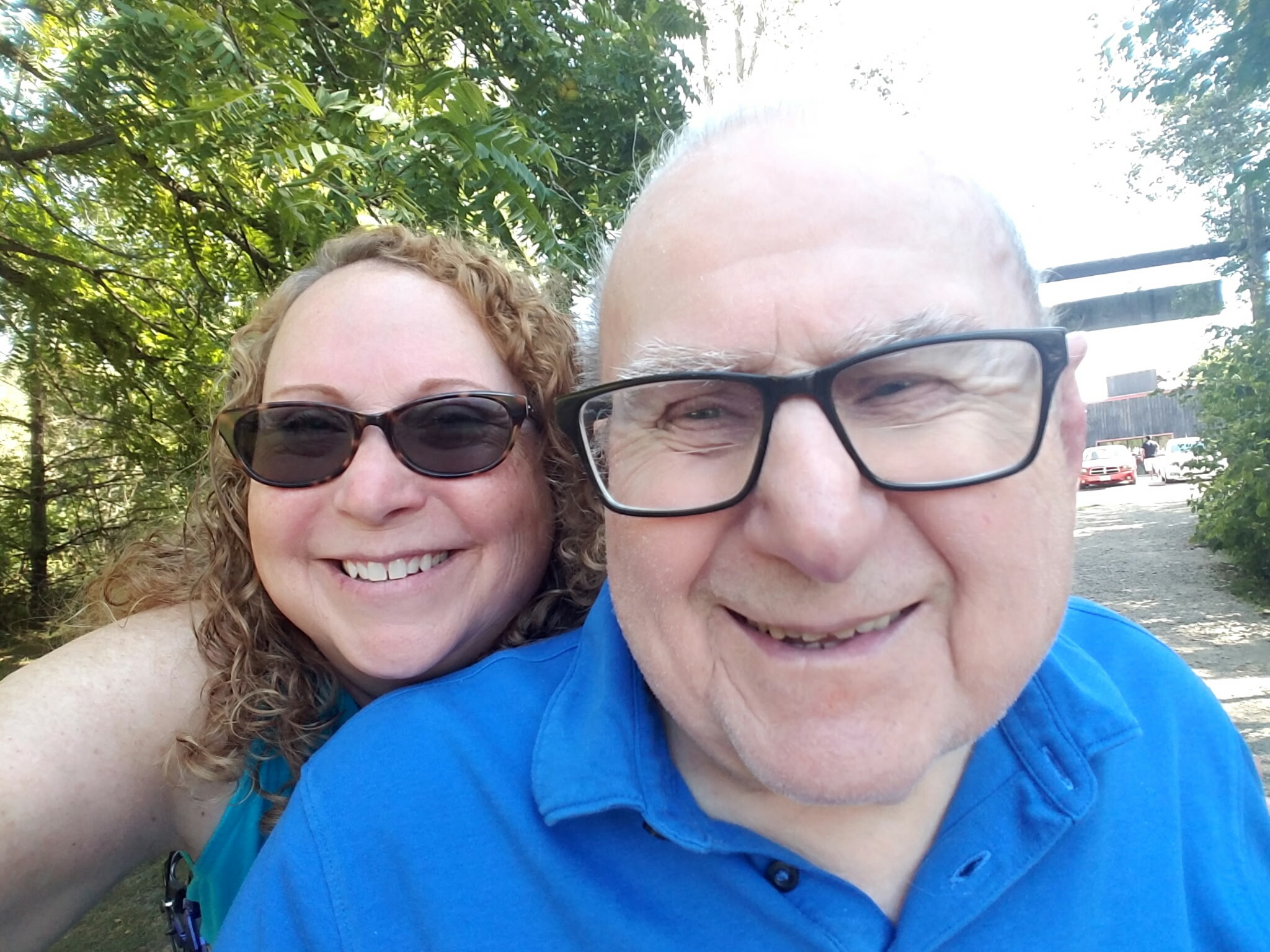 Two people taking a sunny outdoor selfie; a smiling woman with sunglasses beside an older man wearing glasses, both close to the camera with green trees in the background.