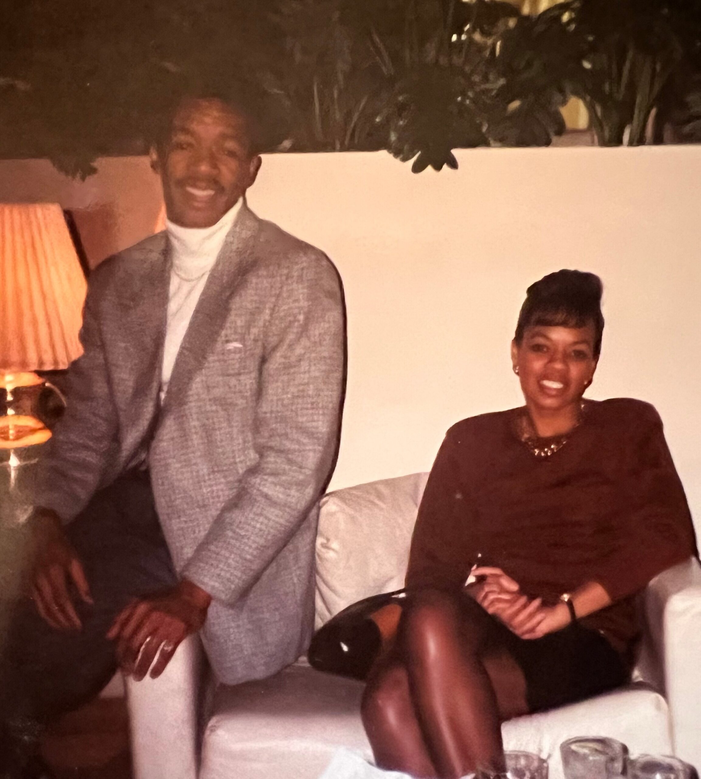 Smiling man in a light gray blazer and white turtleneck beside a smiling woman in a dark dress in a warmly lit room.