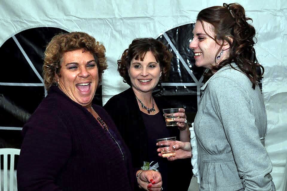 Three women at a party inside a tent, laughing and holding drinks, engaged in conversation.