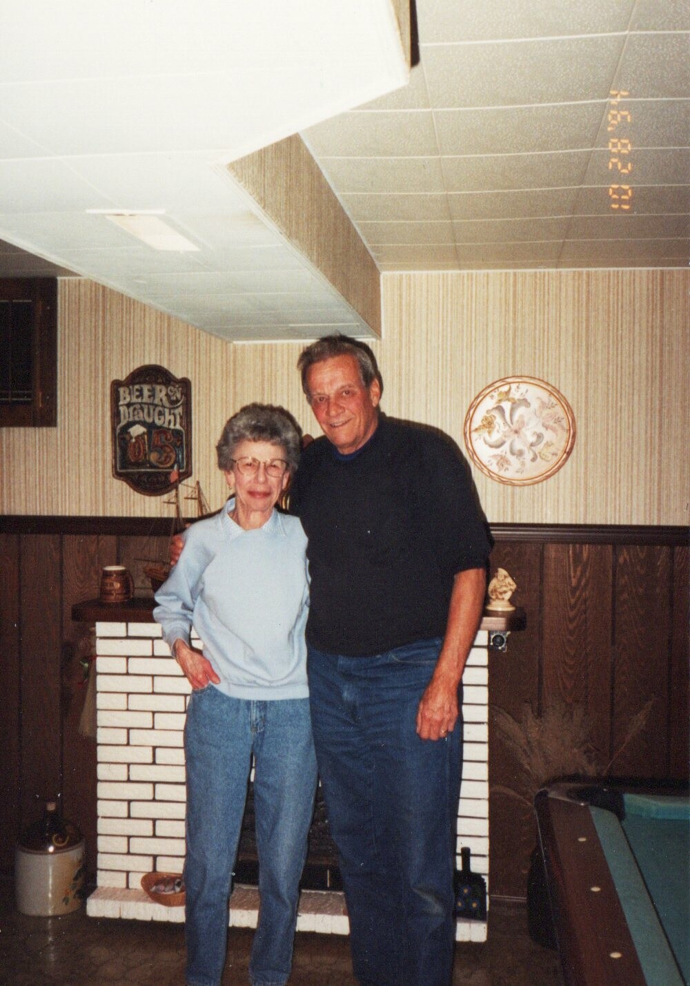 An elderly woman and a man stand arm in arm in a home game room, smiling at the camera by a white brick fireplace.