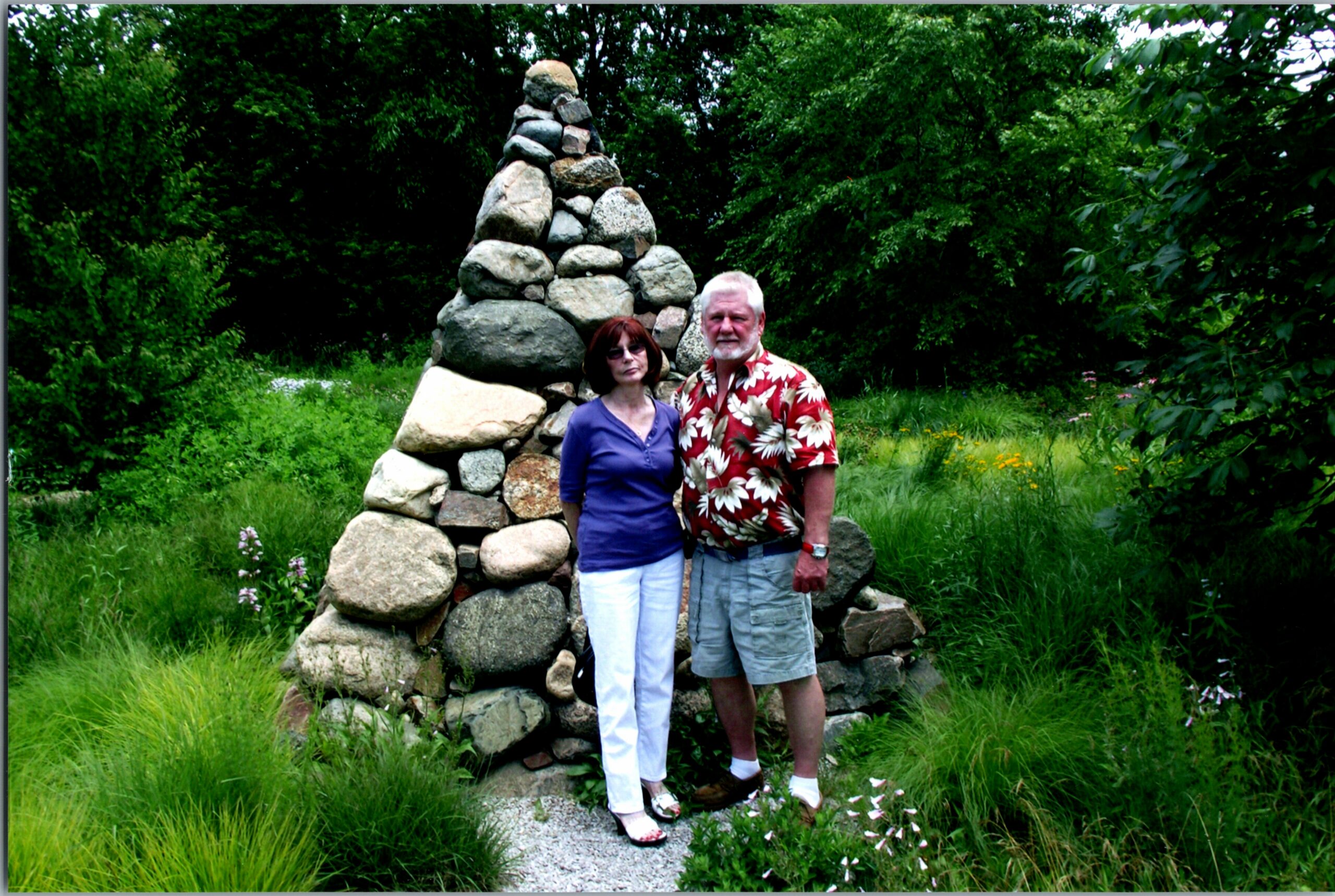 Older couple standing next to a tall stacked-stone cairn in a lush garden.