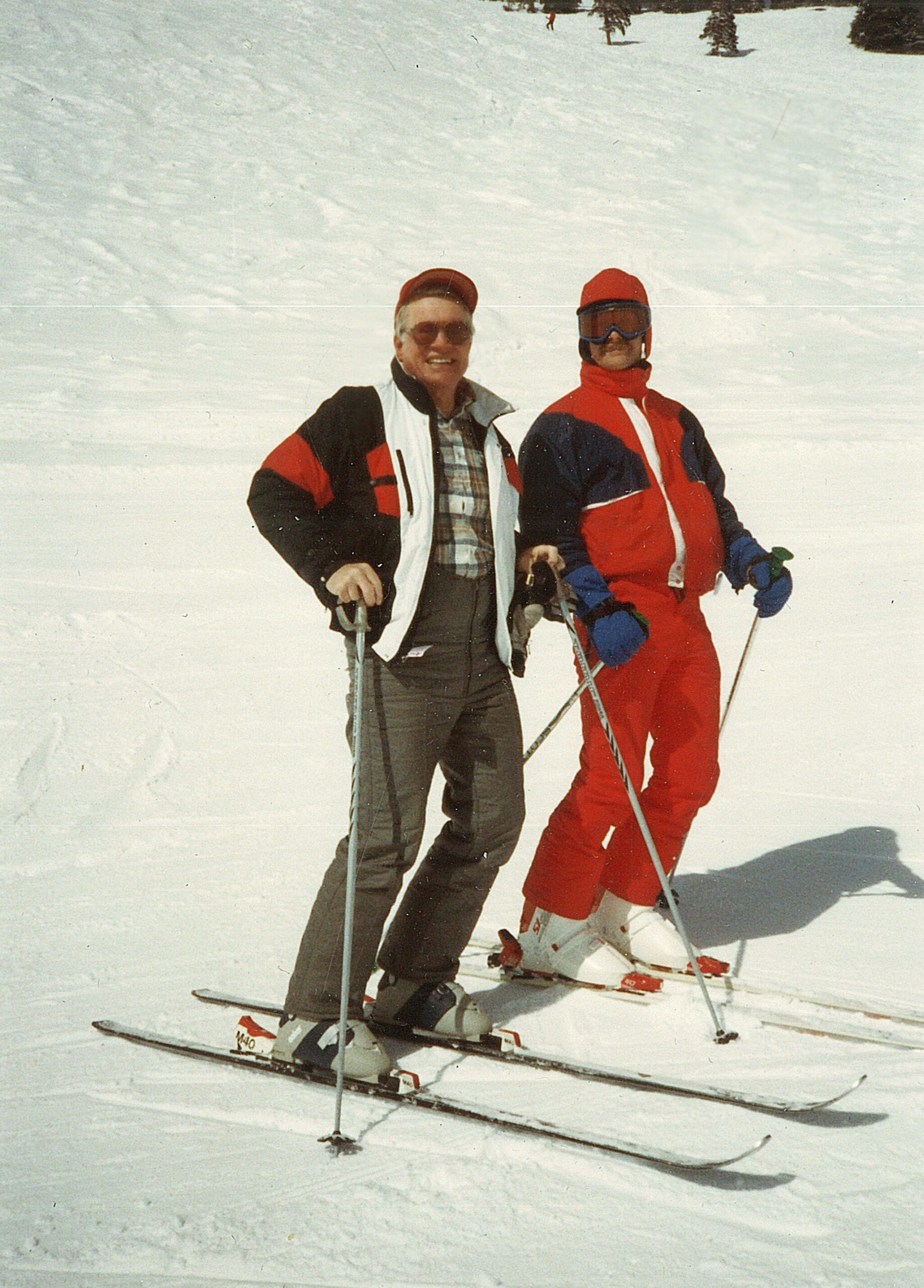 Two skiers pose together in the snow, wearing colorful jackets and goggles, holding ski poles on a sunny slope.