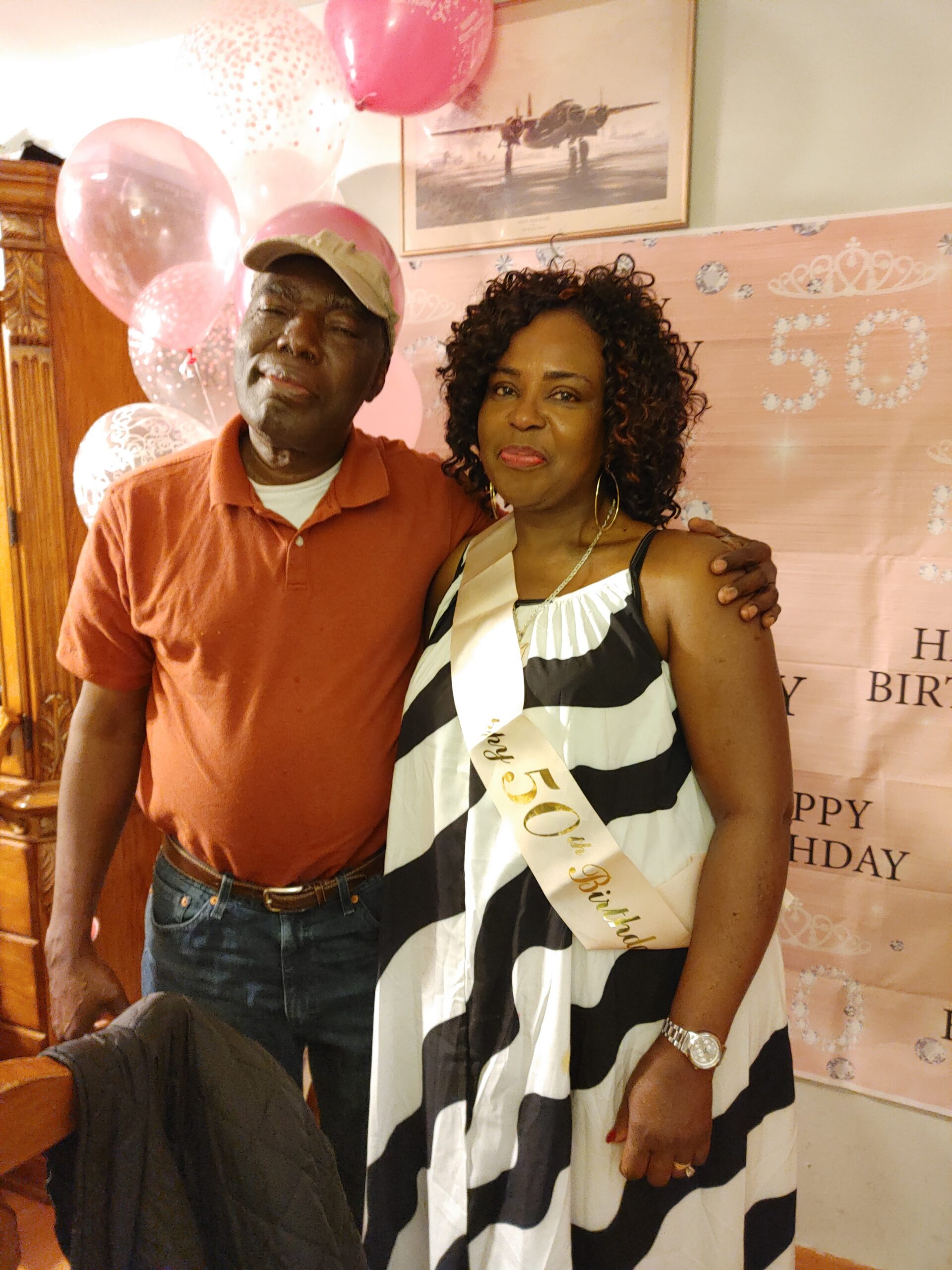 A smiling couple posing together at a birthday party, with pink balloons and a 'Happy Birthday' backdrop behind them.