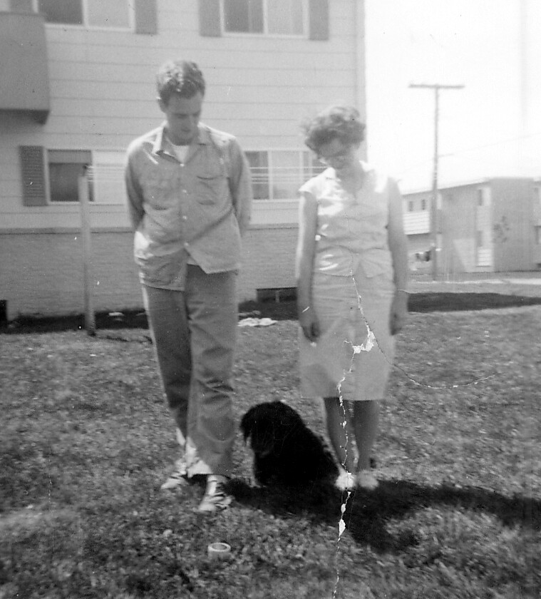 Black-and-white photo of a man and a woman walking a small black dog on a grassy lawn in front of a multiunit apartment building.