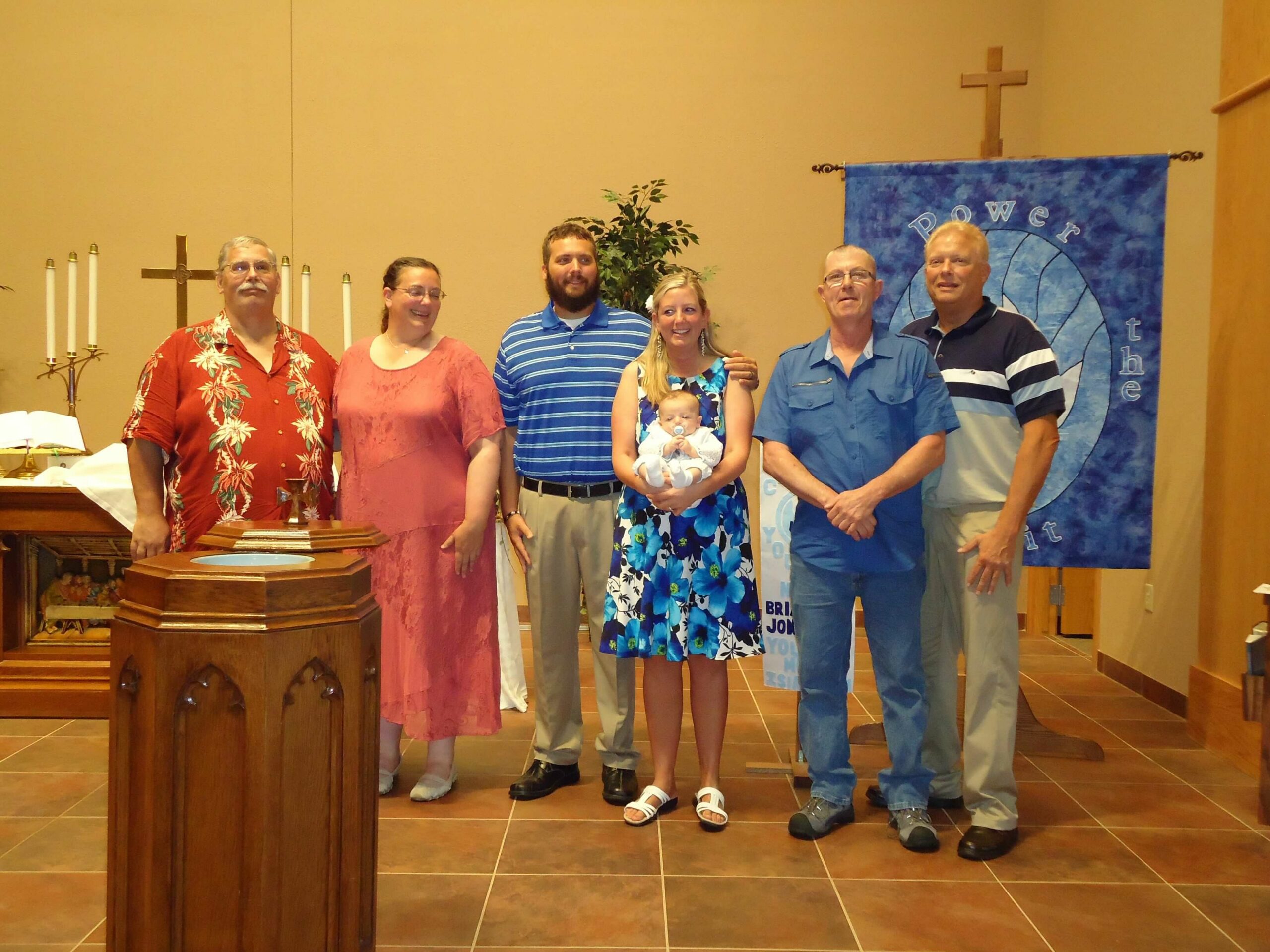 Group of seven people (including a baby) posing on a church stage with a cross and blue banner in the background, smiling for a photo at the front pews and pulpit area.