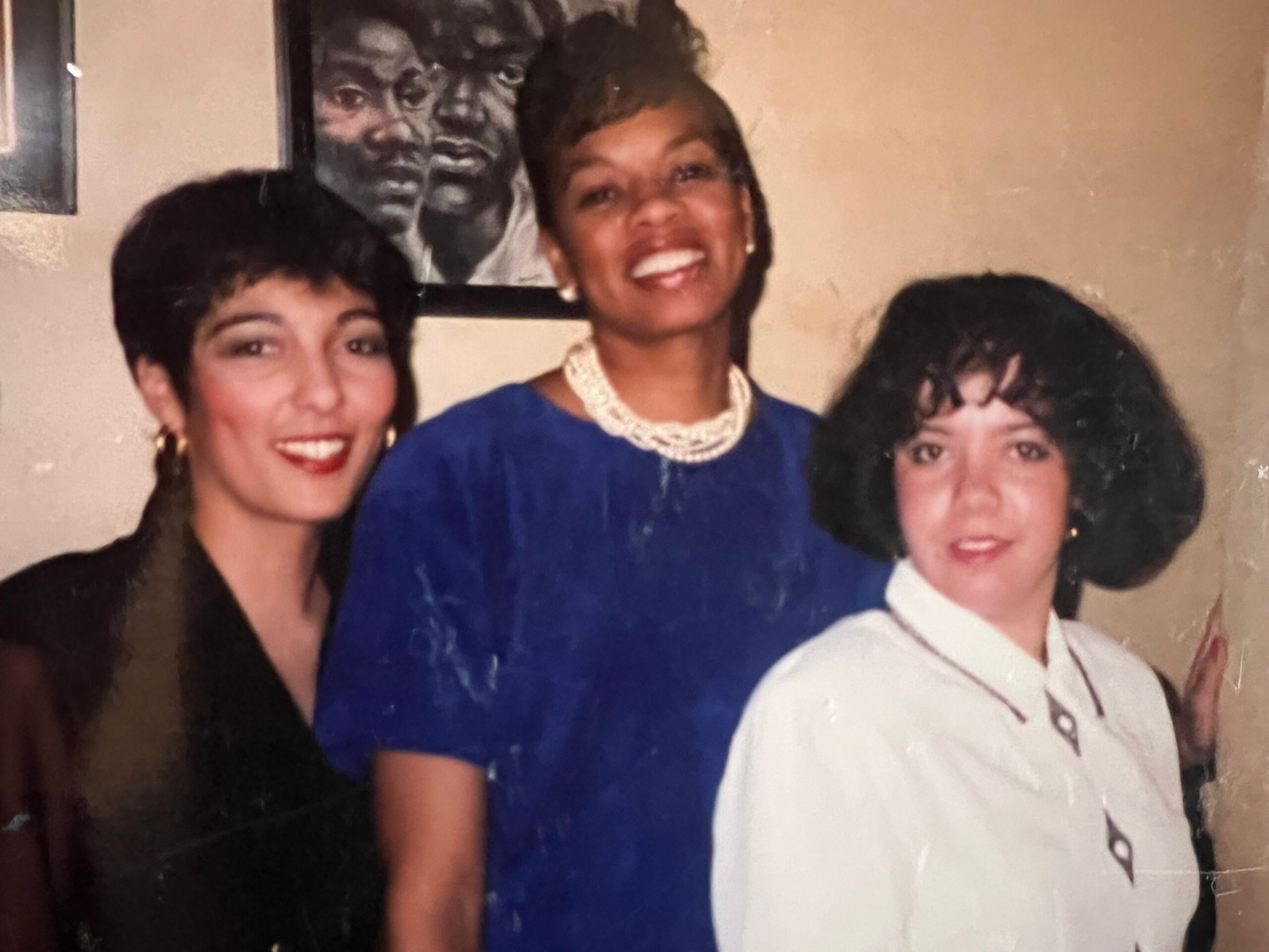 Three smiling women posing together indoors: center woman in a blue dress with a pearl necklace, flanked by women in darker clothing; framed artwork behind them.