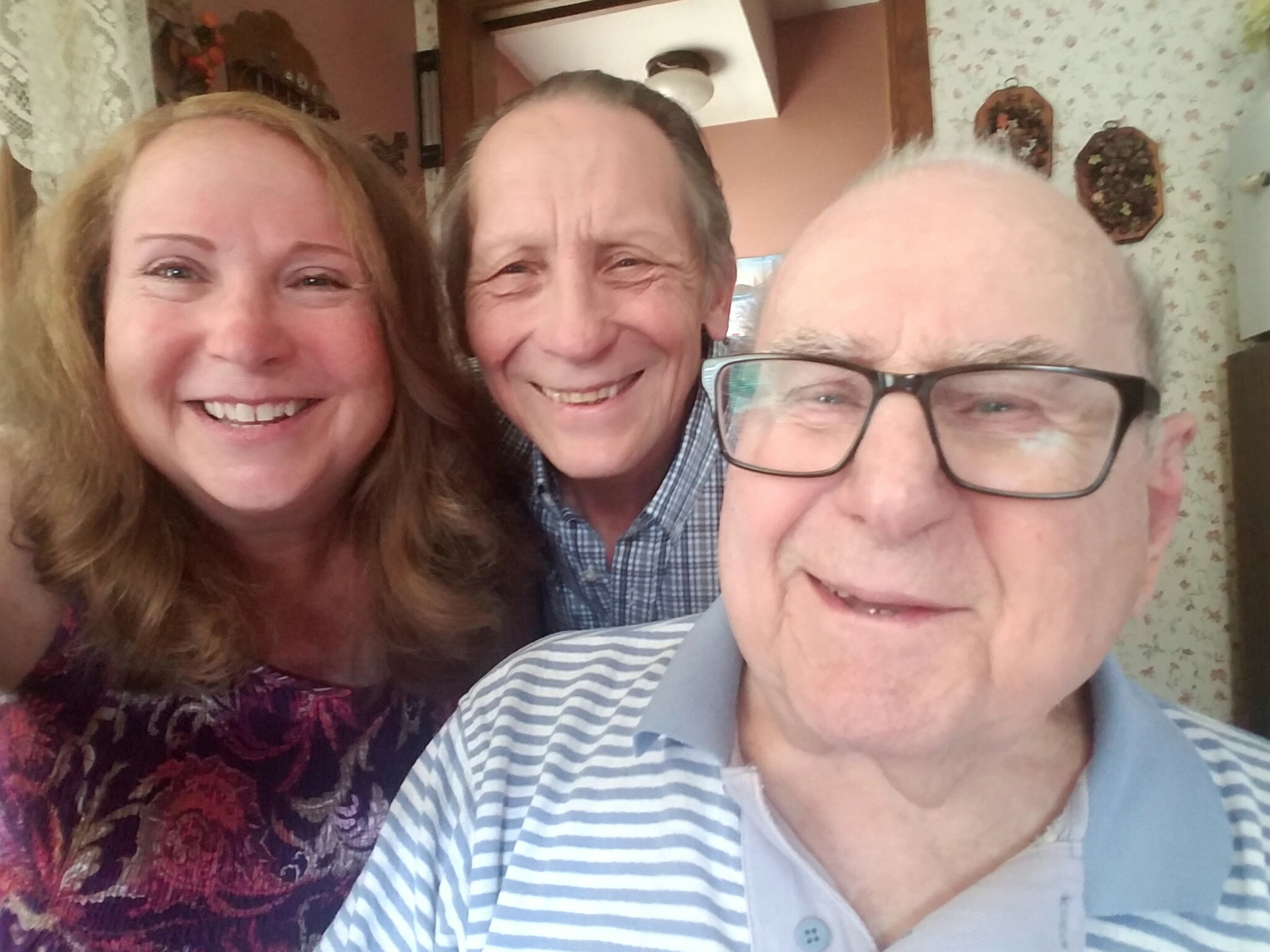 Three adults taking a close-up selfie indoors, smiling at the camera with floral wallpaper in the background (two women and a man).