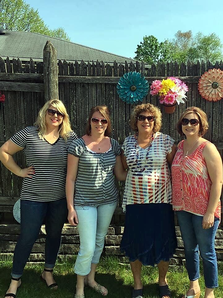 Four women standing side by side outdoors in front of a wooden fence with large colorful paper flowers, smiling at the camera.