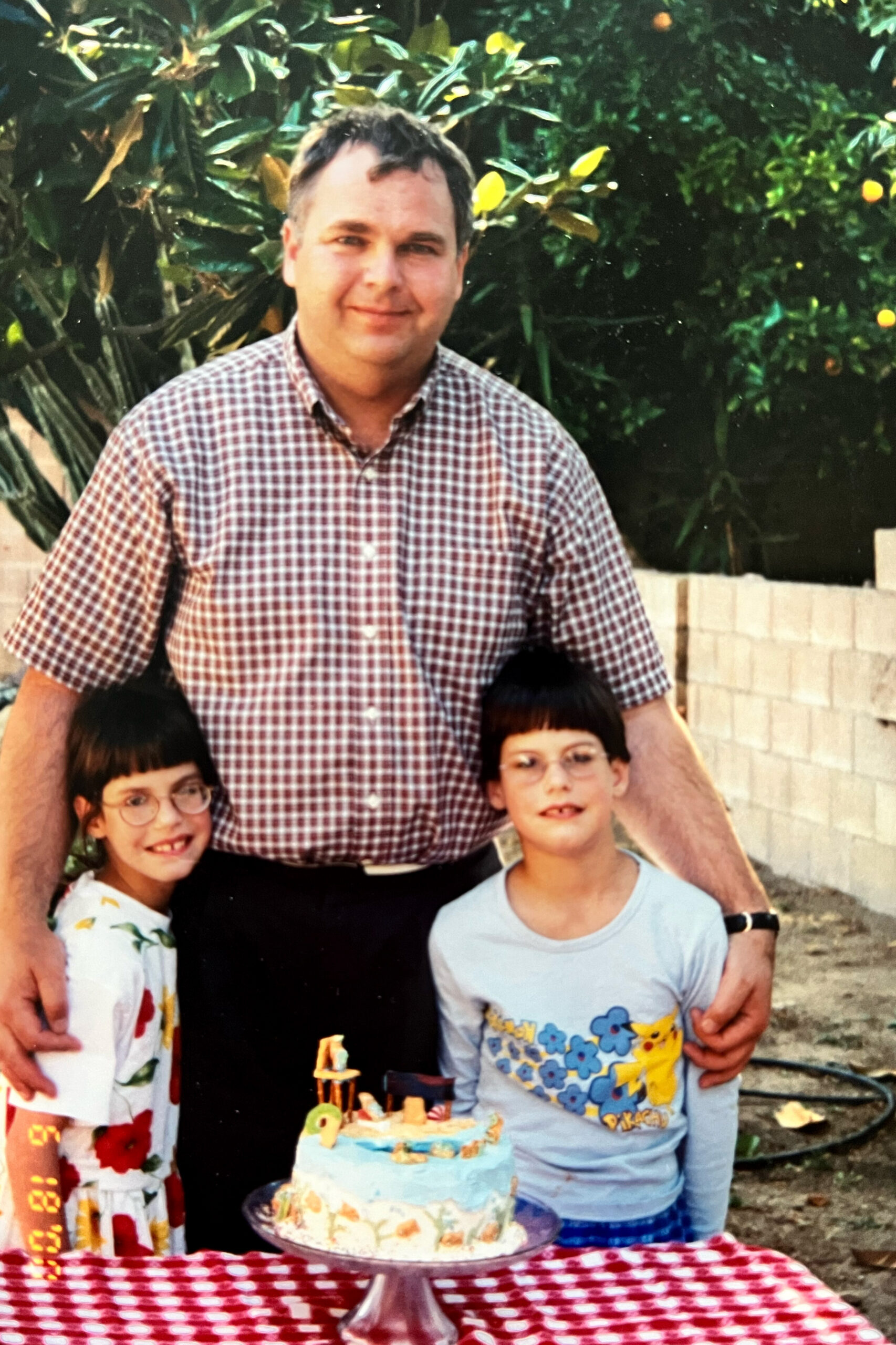 A man in a checkered shirt posing with two children at an outdoor birthday party, with a layered cake on a red-and-white tablecloth in front of them.
