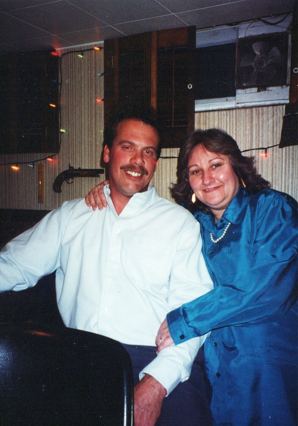 Smiling man in a white shirt sits with his arm around a smiling woman in a blue jacket; warm party lighting in the background.