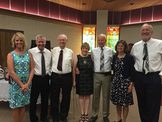 Seven adults in formal attire pose for a group photo in a room with stained-glass windows in the background.