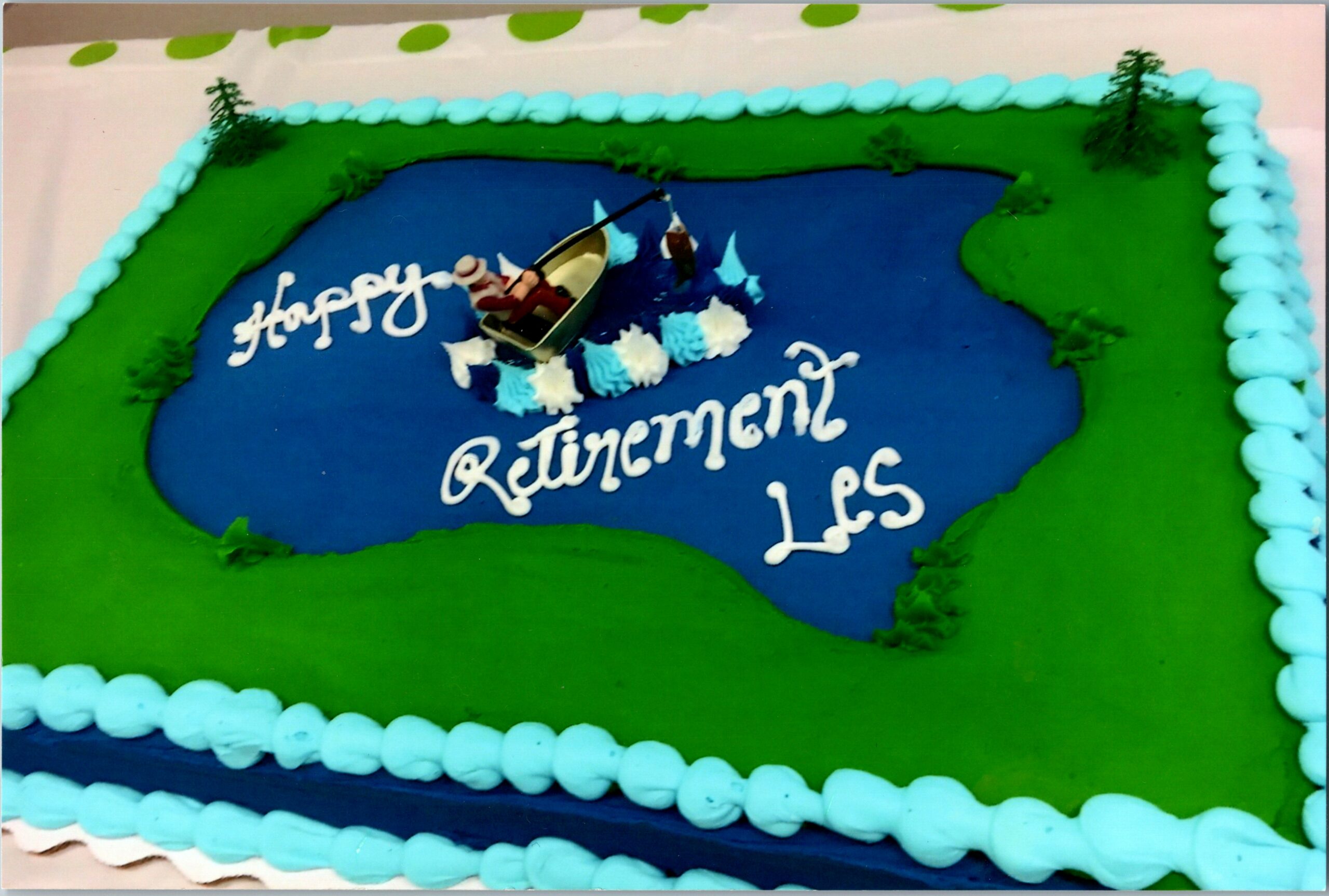 Cake with a blue pond scene, a small boat with passengers, green surrounding land, and 'Happy Retirement' in white icing on blue surface.