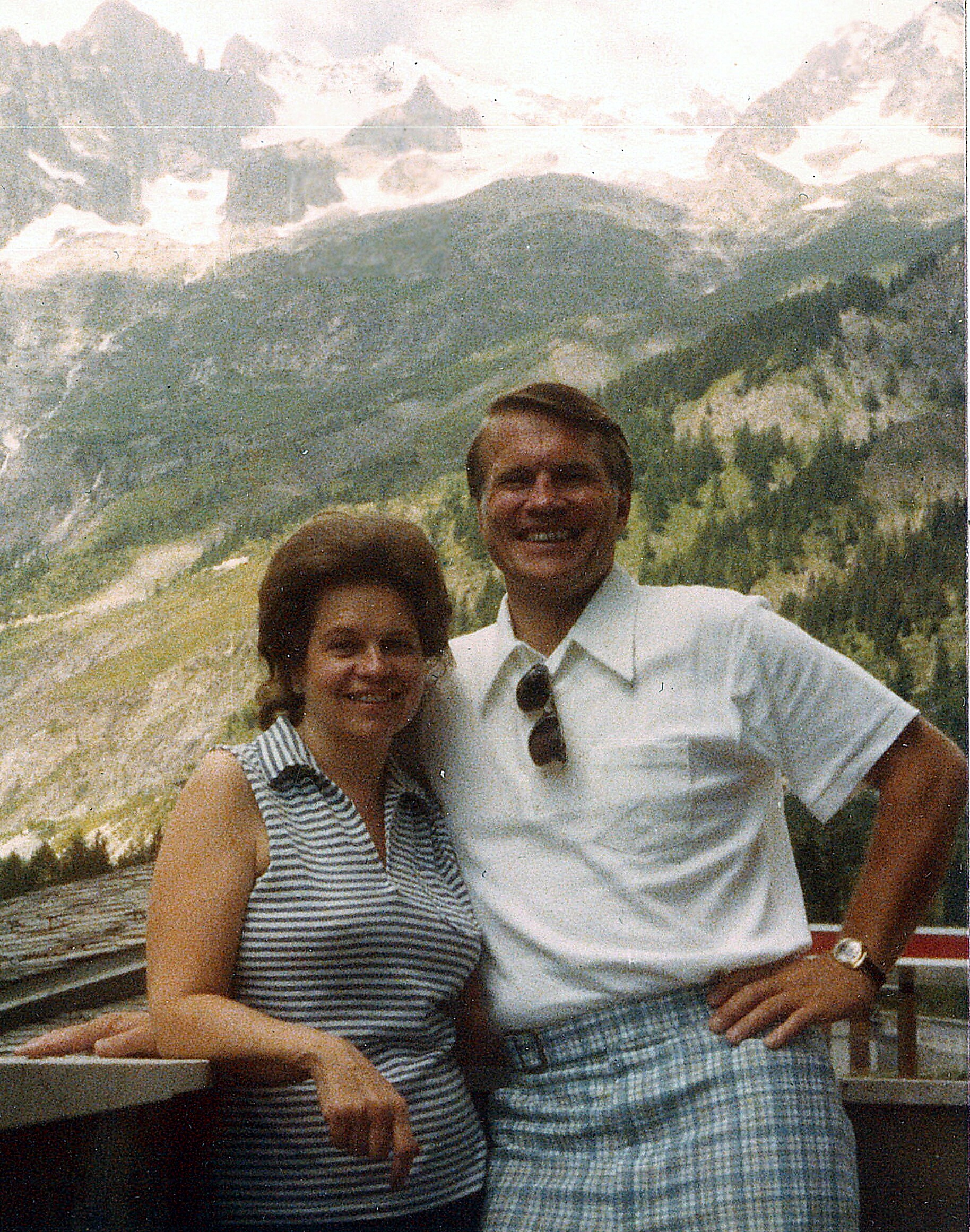 Two people smiling for a photo on a railing with a snowy alpine mountain backdrop behind them.
