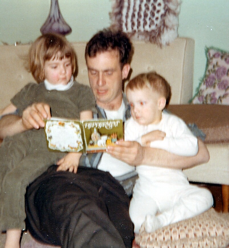 A man sits on a couch reading a children’s book aloud while two kids cuddle close, one on each side.