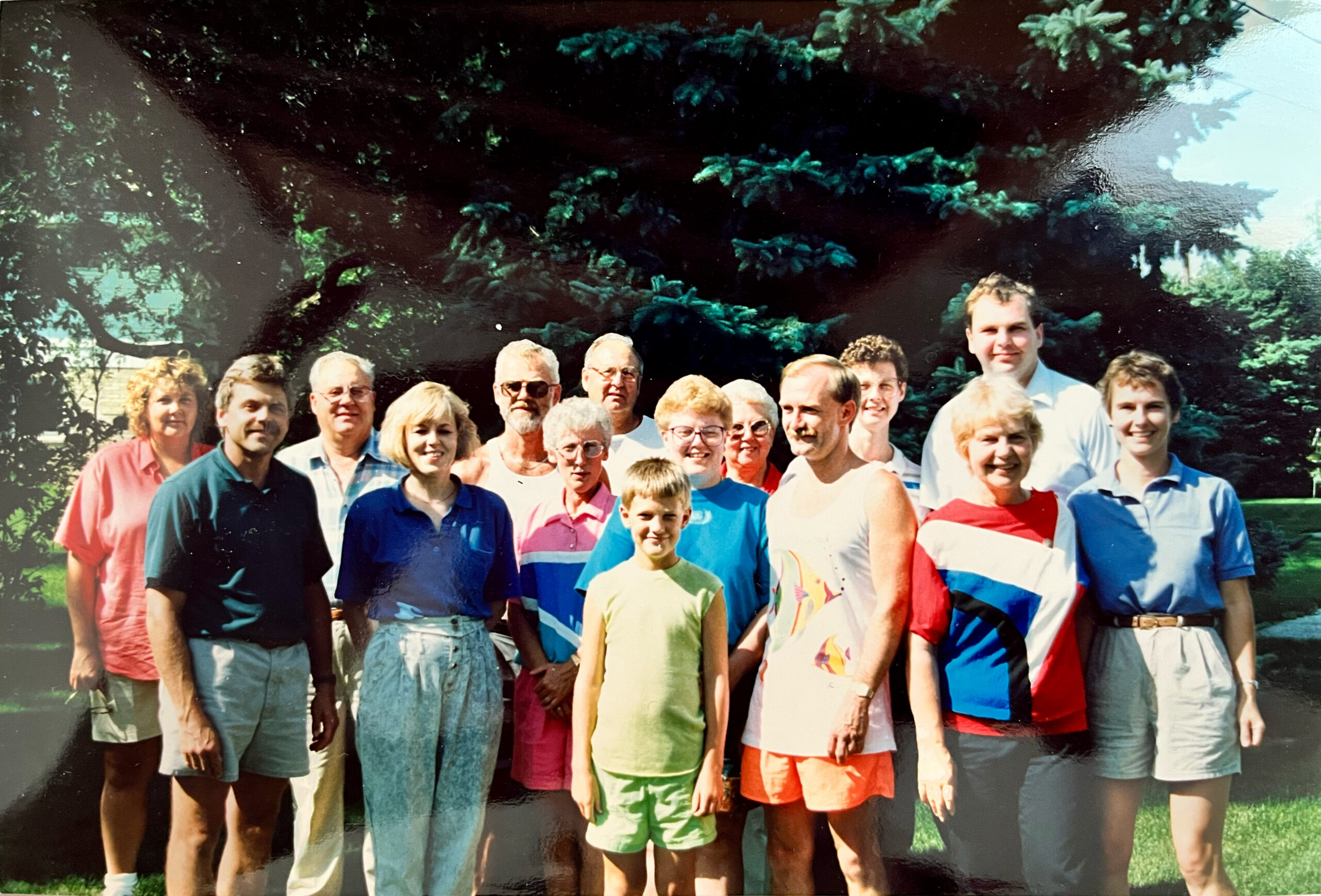 A large group of people of various ages posing together outdoors in a sunny yard with trees in the background, wearing colorful casual clothing.