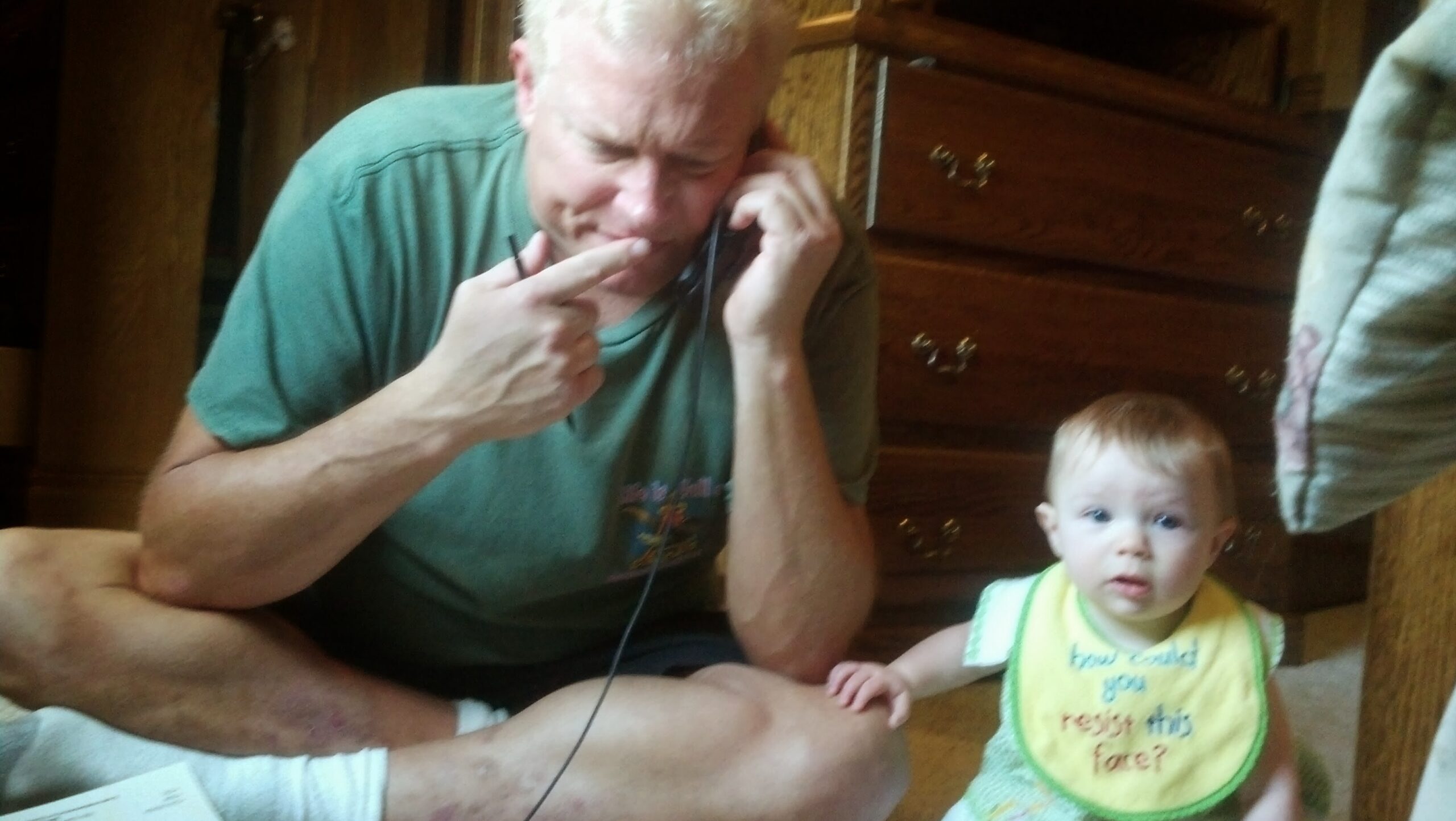 Man in a green shirt talking on a corded phone at a table, with a baby in a yellow bib nearby and wooden furniture in the background.