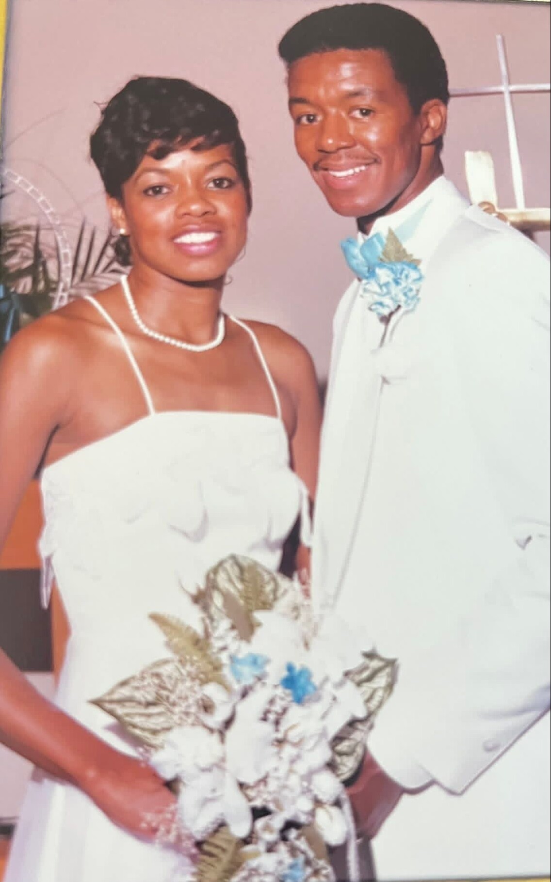 Bride and groom smiling for a wedding photo; she wears a white strapless dress with a pearl necklace and holds a floral bouquet, he wears a light suit with a blue boutonniere/bow tie.