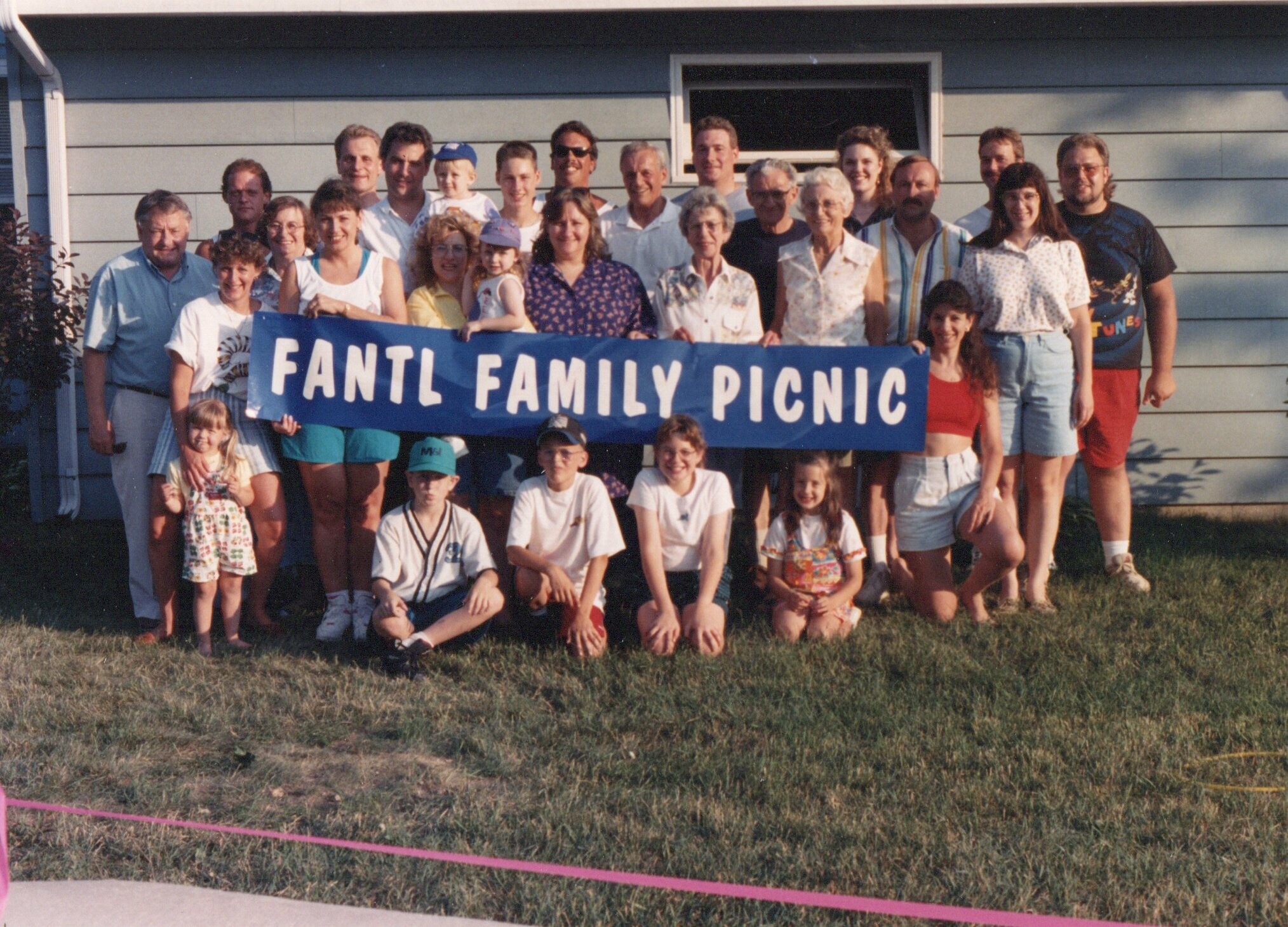 A large group of relatives posing outdoors behind a blue banner that reads 'FANTL FAMILY PICNIC' in a yard.