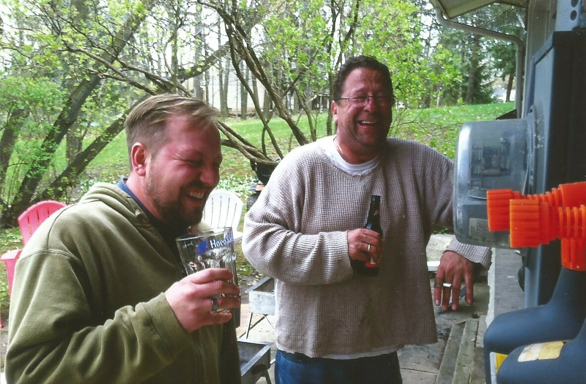 Two men laughing and holding beer bottles outdoors near a beverage dispenser.