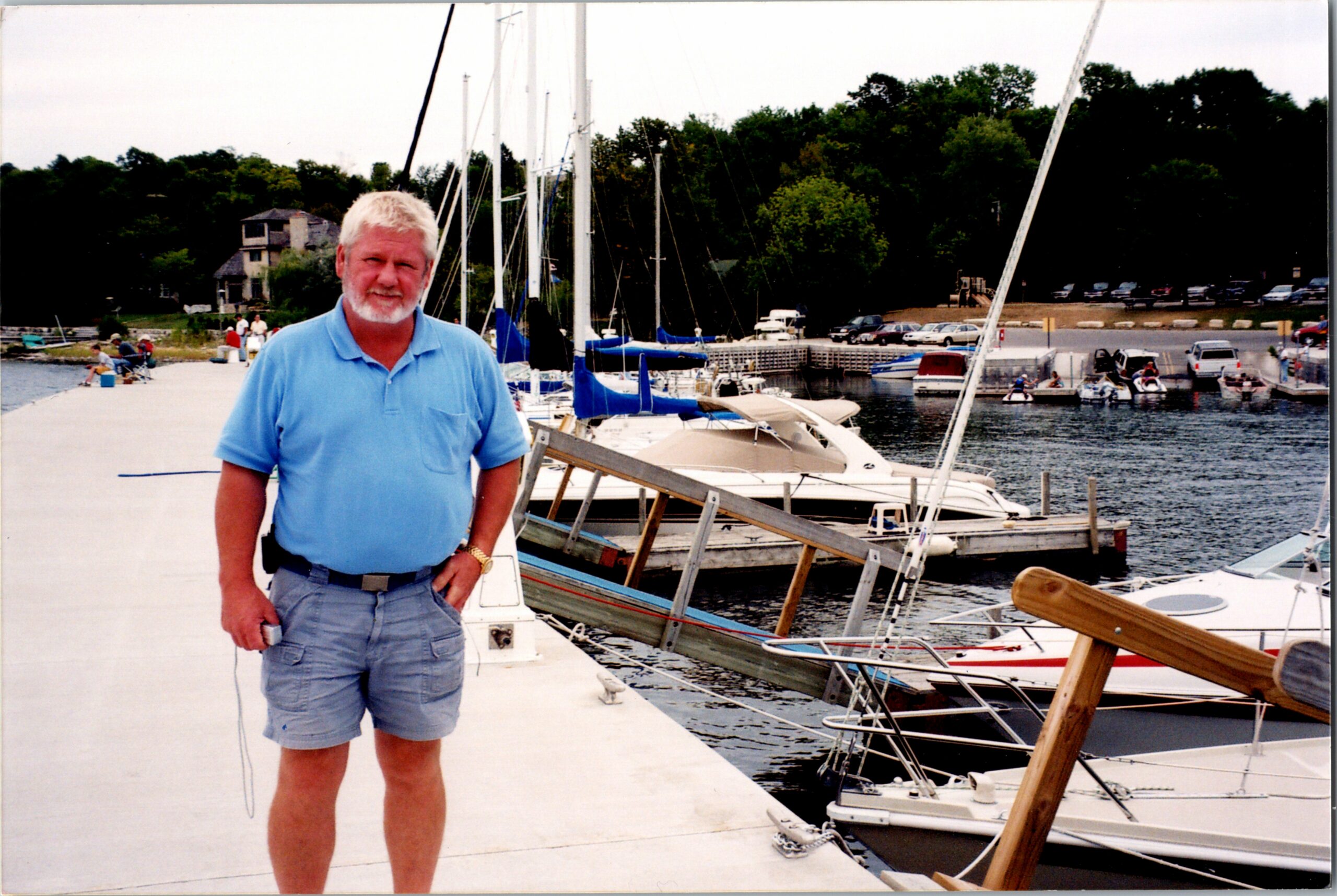 Older man with white hair and beard in a light blue polo standing on a dock near moored sailboats at a marina