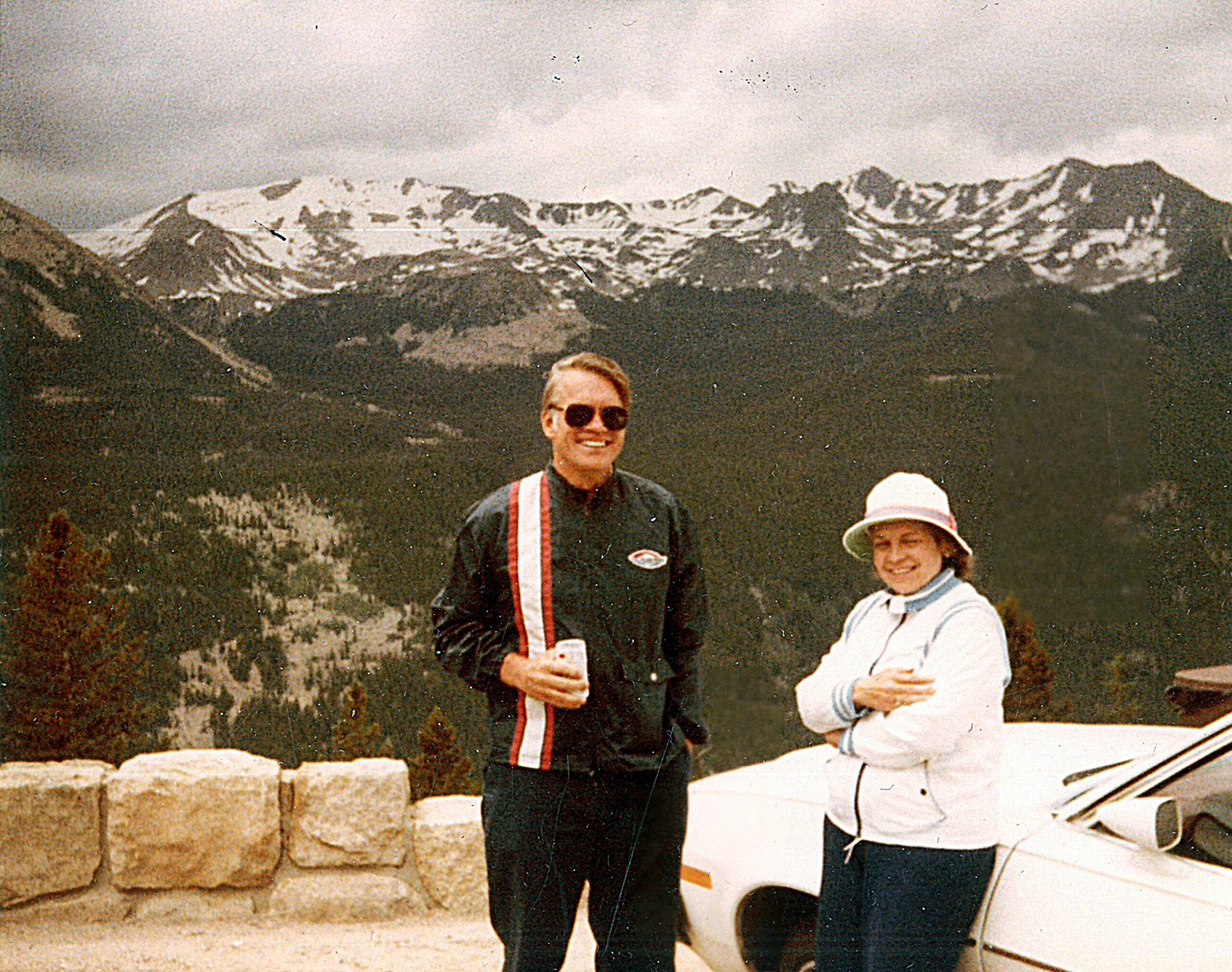 Two people posing outdoors with a snow-capped mountain range in the background; man wearing dark jacket and sunglasses, woman in a white jacket and hat.