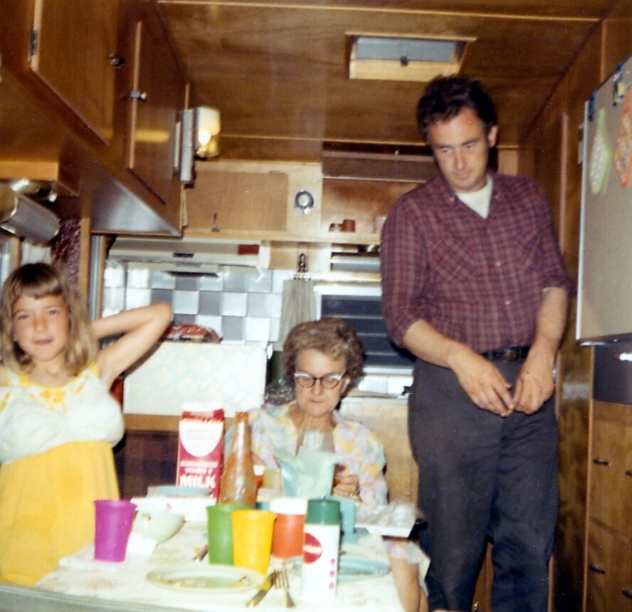 A family of three in a retro kitchen around a cluttered table: a girl in a yellow dress on the left, an elderly woman with glasses at the table, and a man in a plaid shirt standing on the right.