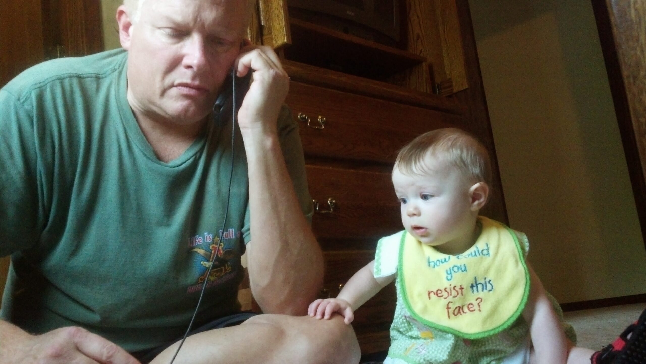 Man in a green T-shirt talks on a corded phone while a baby in a yellow bib sits nearby, reaching toward him in a room with wooden furniture behind.
