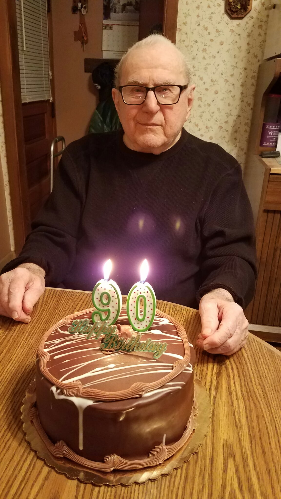 Elderly man with glasses sits at a table behind a chocolate birthday cake with two lit candles forming '90'