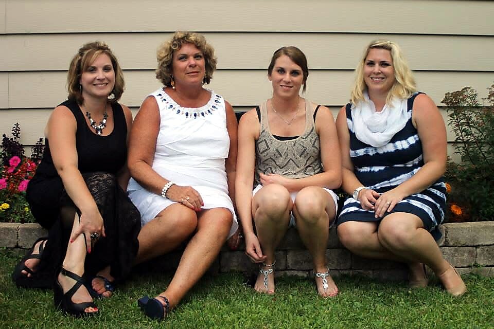 Four women sit on a low garden wall, posing and smiling for the camera; from left: black-dressed woman, woman in white dress, young woman in a patterned tank top and shorts, and a woman in a blue‑and‑white striped dress with a white scarf.